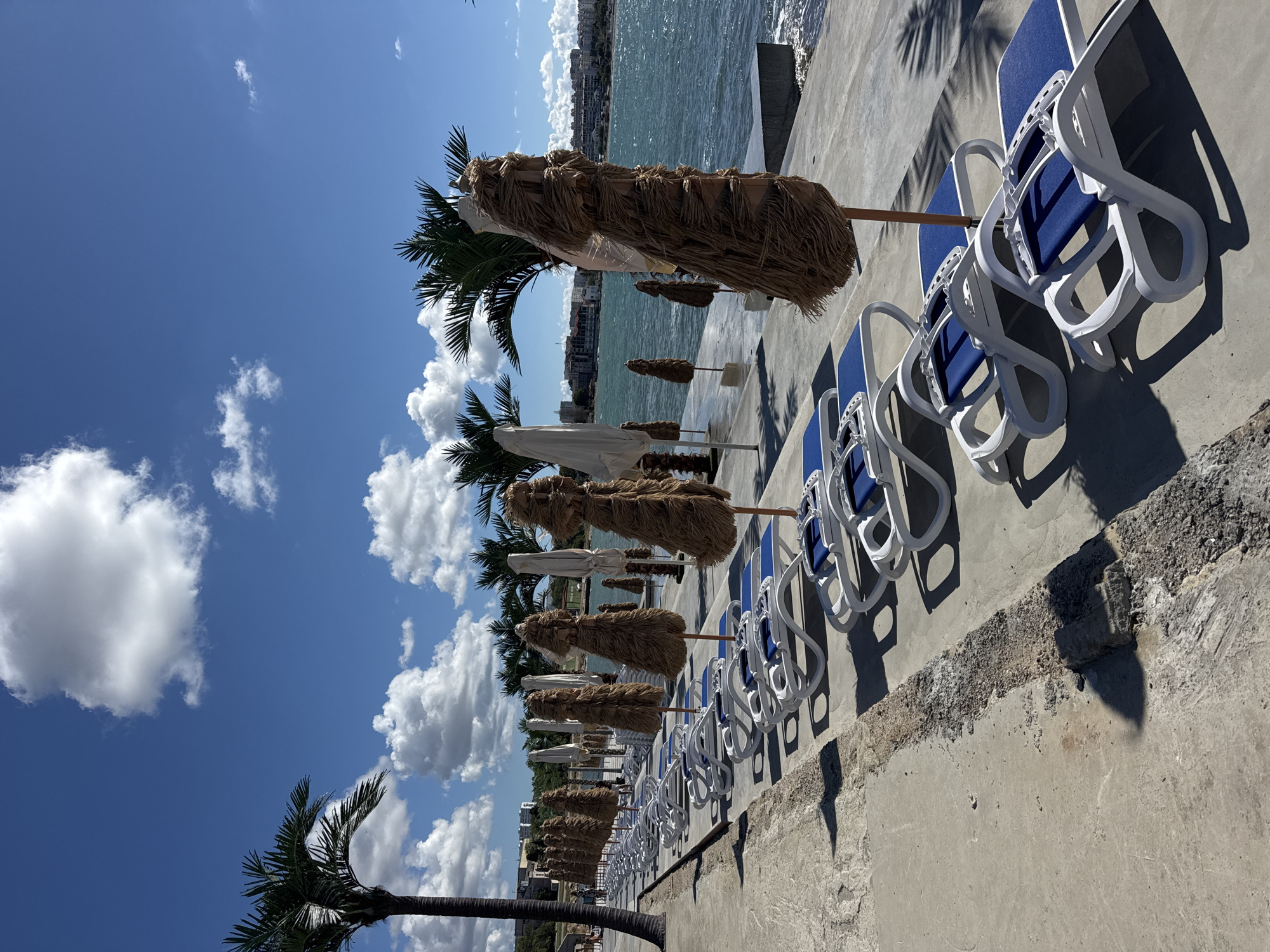 The image depicts a beachside promenade in Sevastopol, Ukraine, on a sunny day with scattered clouds. A row of white plastic lounge chairs with blue cushions is lined up on a concrete walkway. Some of the chairs are covered with protective plastic sheeting. Behind the chairs, a line of thatched umbrellas stands, most of them also covered for protection. Palm trees are visible, with one tall palm on the left and others further down the promenade. The sea is to the right, with a gentle wave pattern and a visible shoreline with buildings in the distance. The concrete walkway shows signs of wear and tear in some sections, with exposed aggregate. A person can be seen walking in the background on the left side of the promenade.