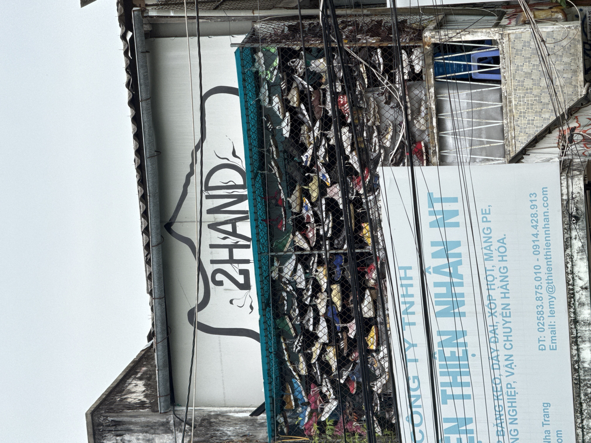 The image shows a building facade in Nha Trang, Vietnam, with a large sign that reads "2HAND". Below the sign, a mesh fence is filled with numerous pairs of athletic shoes, suggesting a shoe resale or secondhand shop. Electrical wires are prominently visible in the foreground, crossing in front of the building and the shoe display. A smaller sign below the "2HAND" sign contains Vietnamese text and contact information, including a phone number and email address, with "Nha Trang" mentioned. The sky is overcast. The building appears weathered, with peeling paint and visible signs of age.