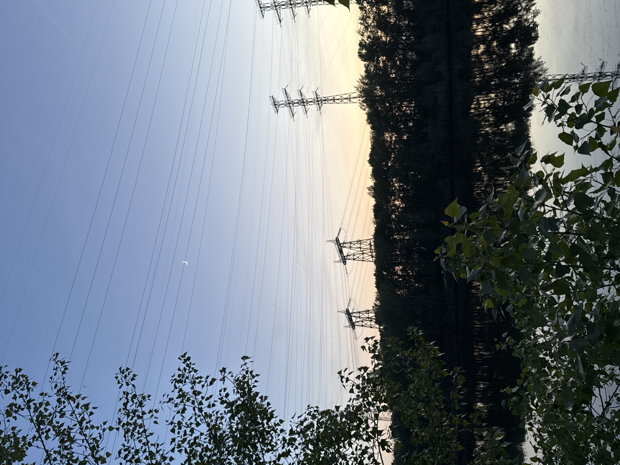 The image, likely taken in Chaikovsky, Russia, is oriented sideways, with the sky to the left. Power lines stretch diagonally across the frame. Steel transmission towers punctuate the line's path. A sliver of a crescent moon is visible in the upper left quadrant. Tree foliage occupies the lower portion of the image. Reflected trees create a mirror-like effect on the right.