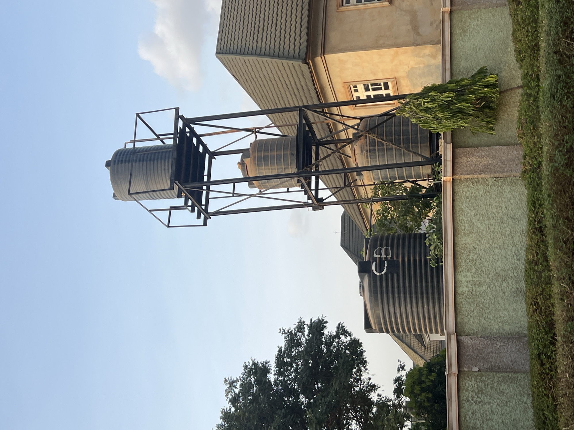 The image, rotated 90 degrees clockwise, captures an outdoor scene in Abuja, Nigeria, under a bright, clear blue sky with a few faint clouds. The prominent subjects are several black water tanks and a building.
In the left and central parts of the frame, a tall, black metal support structure holds two large, cylindrical black water tanks high above the ground. Another large, ribbed black water tank is visible at ground level towards the right. This ground-level tank features white markings on its side, which appear to read "C U" or "G U" possibly with additional symbols.
Behind the elevated water tanks, a multi-story building stands with light brown stucco walls, white-framed windows, and a dark grey, ridged roof. Green foliage, including a tall, leafy tree in the lower center and a manicured hedge running along the right side of the image, indicates a verdant setting. The scene is bathed in daylight, suggesting either morning or afternoon. No specific activity or event is taking place; it's a static view of the infrastructure.