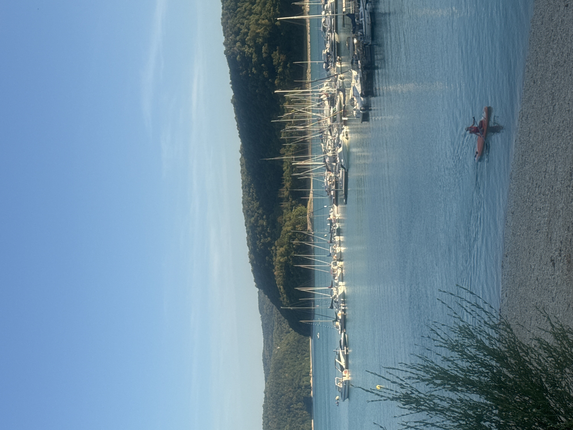 The image depicts a tranquil harbor scene under a clear blue sky. Several sailboats and smaller motorboats are moored in the calm, turquoise water. A lone individual is paddling an orange stand-up paddleboard in the foreground, leaving a gentle wake. In the background, lush, green, tree-covered hills rise from the water's edge, creating a scenic backdrop. The shore in the immediate foreground is composed of pebbles. This scene is located in the area of the lake of Vouglans, near the town of Maisod, France.
