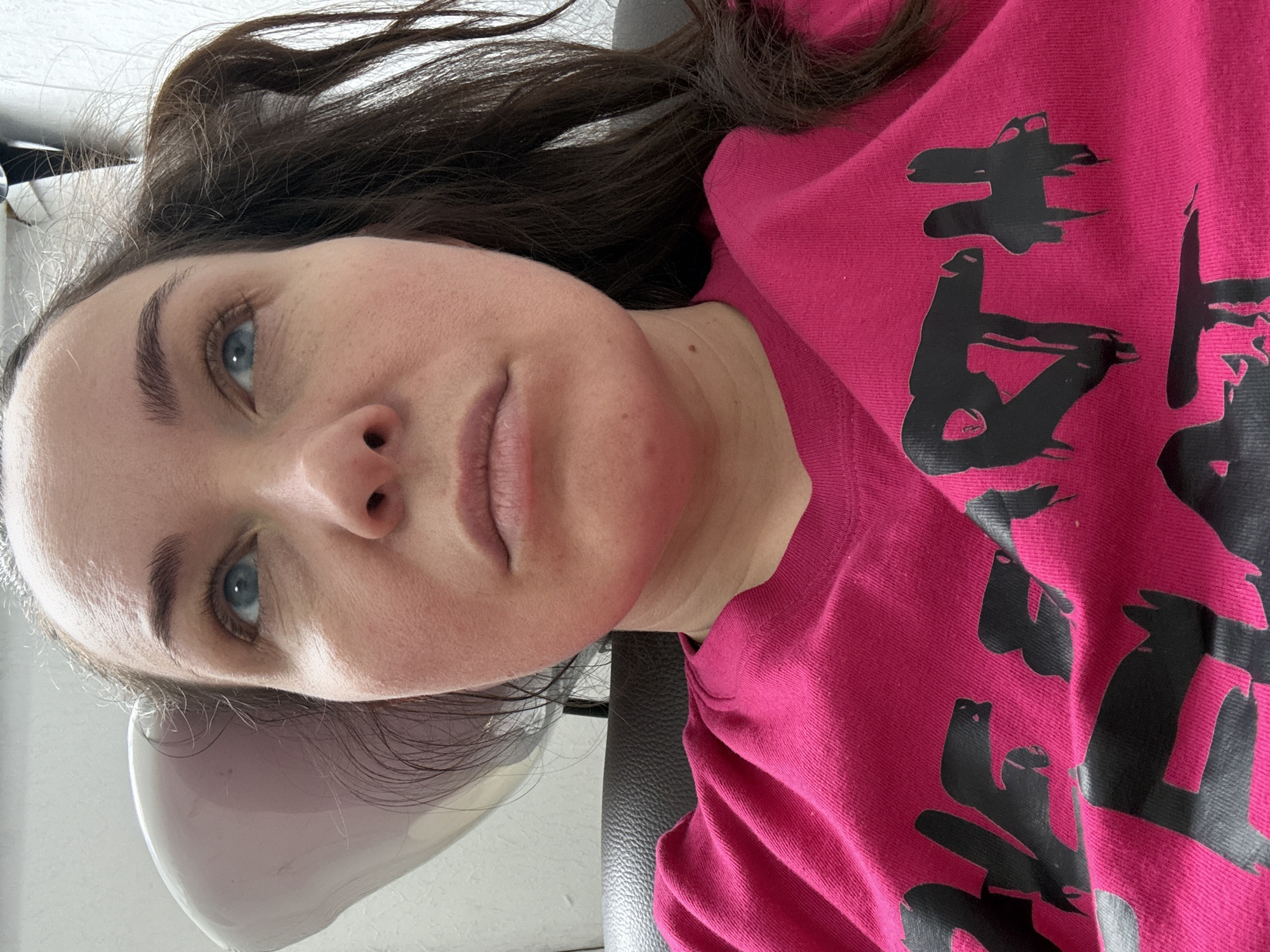 The media file presents a close-up portrait of a woman, oriented sideways (rotated 90 degrees counter-clockwise from an upright position). She has long dark brown hair and bright blue eyes, looking upwards with a neutral expression. She is wearing a bright pink t-shirt featuring large, black, stylized graphics across the chest. These graphics resemble abstract symbols or characters, possibly an interpretation of Asian script or modern art, with one detail in a cropped view suggesting a cat-like figure.
The woman is lying down, with her head resting on a light-colored, curved headrest or basin, typical of a salon environment. A darker, textured material, likely the upholstery of a chair or salon equipment, is visible behind her head. There is noticeable redness on her cheeks and extending down her neck area. The scene takes place indoors, under what appears to be ambient lighting, consistent with a salon or similar facility in Chaikovsky, Russia. No specific time of day or weather conditions are discernible.