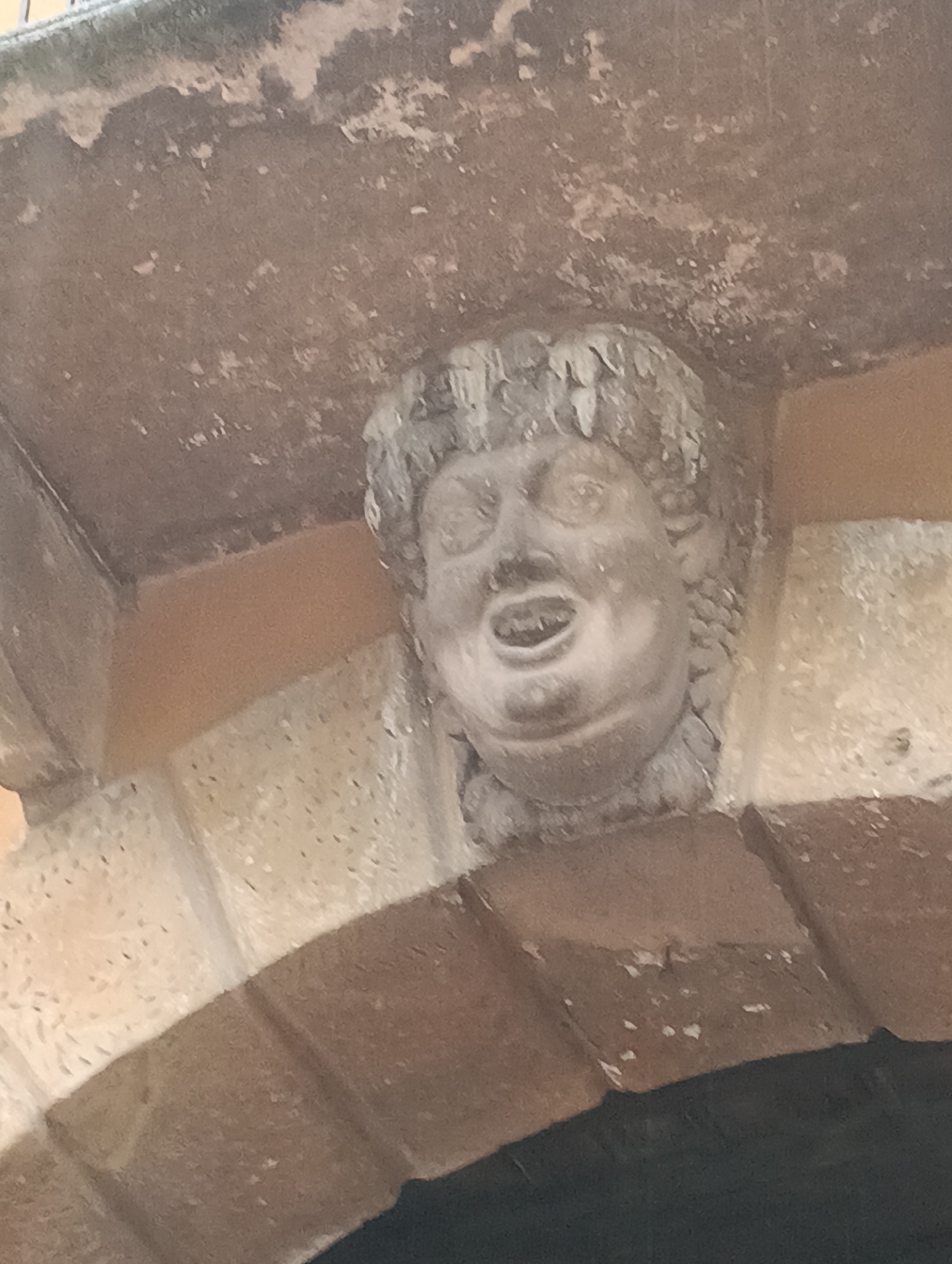 A weathered stone grotesque face, possibly a gargoyle or corbel, is carved into the underside of an archway. The face has a wide open mouth with visible teeth, bulging eyes, and curly hair. It is located on the facade of a building in Verona, Italy, appearing to be part of a historical structure. The surrounding architecture is made of stone and stucco, showing signs of age and wear.