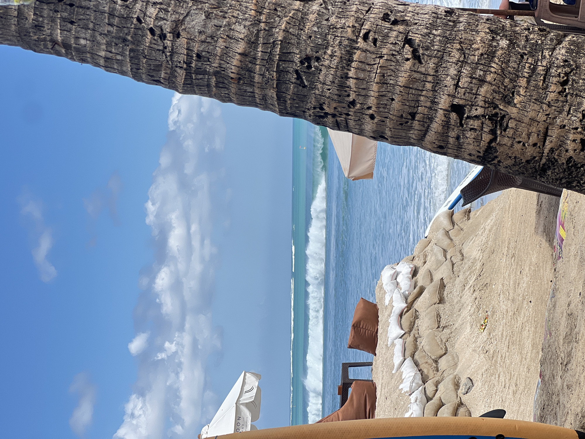 This image depicts a beach scene in Kuta, Indonesia, under a bright, partly cloudy sky. In the foreground, a large palm tree trunk is prominently visible on the right side, its rough bark textured and dark. To the left, a portion of an orange and blue surfboard stands upright.
Further back, a white beach umbrella, with the word "GOON" visible on it, provides shade over a low table and two brown beanbag chairs situated on a sandy embankment. This embankment is reinforced with sandbags. Beyond this seating area, the turquoise ocean stretches out, with a series of powerful, white-capped waves rolling towards the shore. In the distance, the horizon is a clear line where the ocean meets the sky. Another beach umbrella, this one a light tan color, is partially visible between the palm tree and the ocean.
The scene is peaceful and suggests a popular tourist spot, likely a resort or beach club, designed for relaxation and enjoying the ocean. There are no people clearly visible in the frame, though the presence of seating and umbrellas implies recent or imminent human activity. The overall environment is tropical, characterized by the palm tree, sandy beach, and ocean.