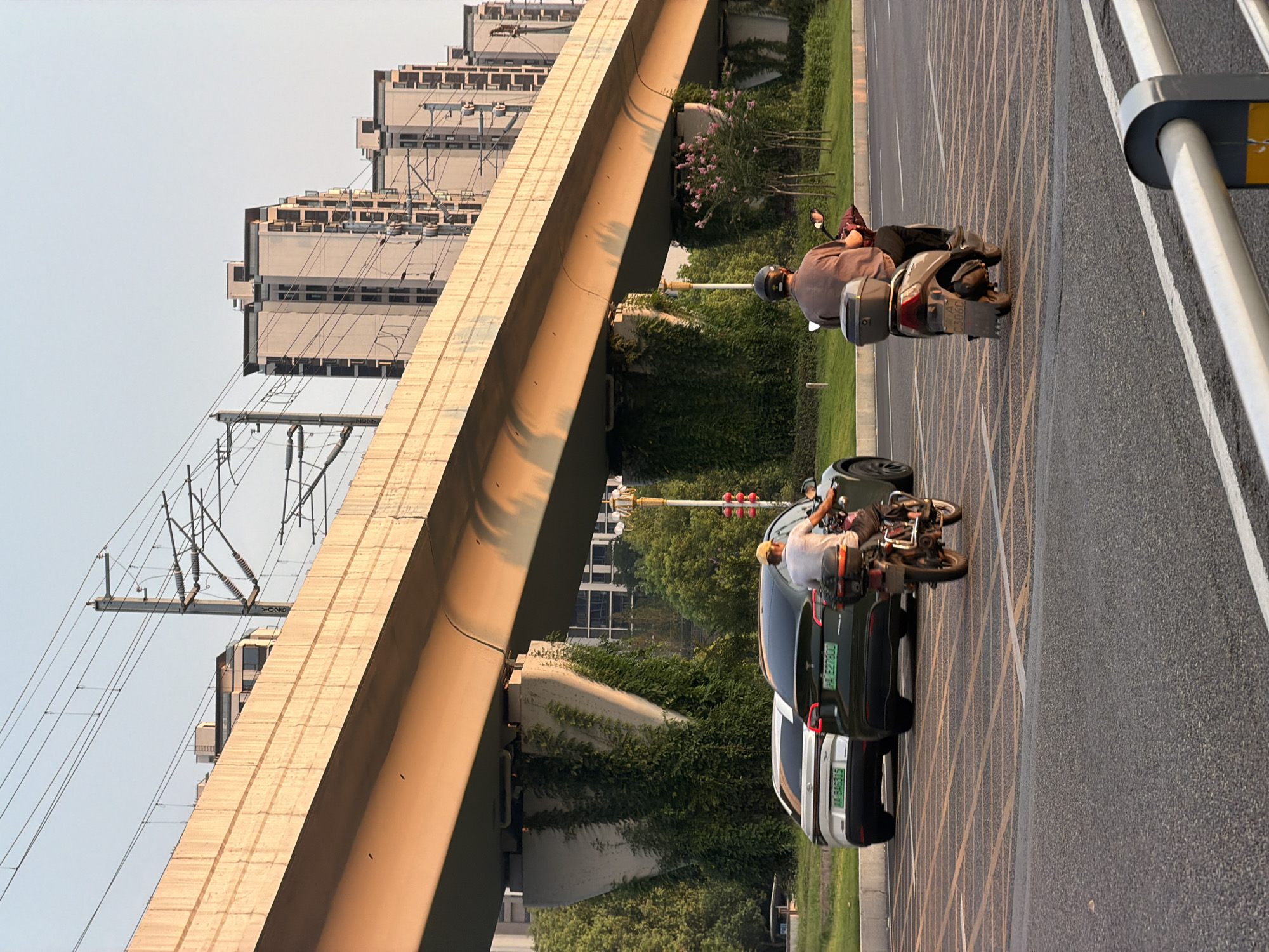 In Jiangjin, China, an overhead elevated railway structure casts a long shadow across a paved road. Two individuals are riding on scooters. The first scooter is ridden by a person wearing a yellow cap and a grey shirt, moving from left to right. Behind them, a second scooter is occupied by a person in a grey shirt and black pants, also traveling from left to right. To the left of the road, two cars are parked, a white SUV and a dark green Tesla. The background features residential buildings and lush green trees, with wires from the elevated structure crisscrossing the sky. The lighting suggests it is late afternoon or early evening.