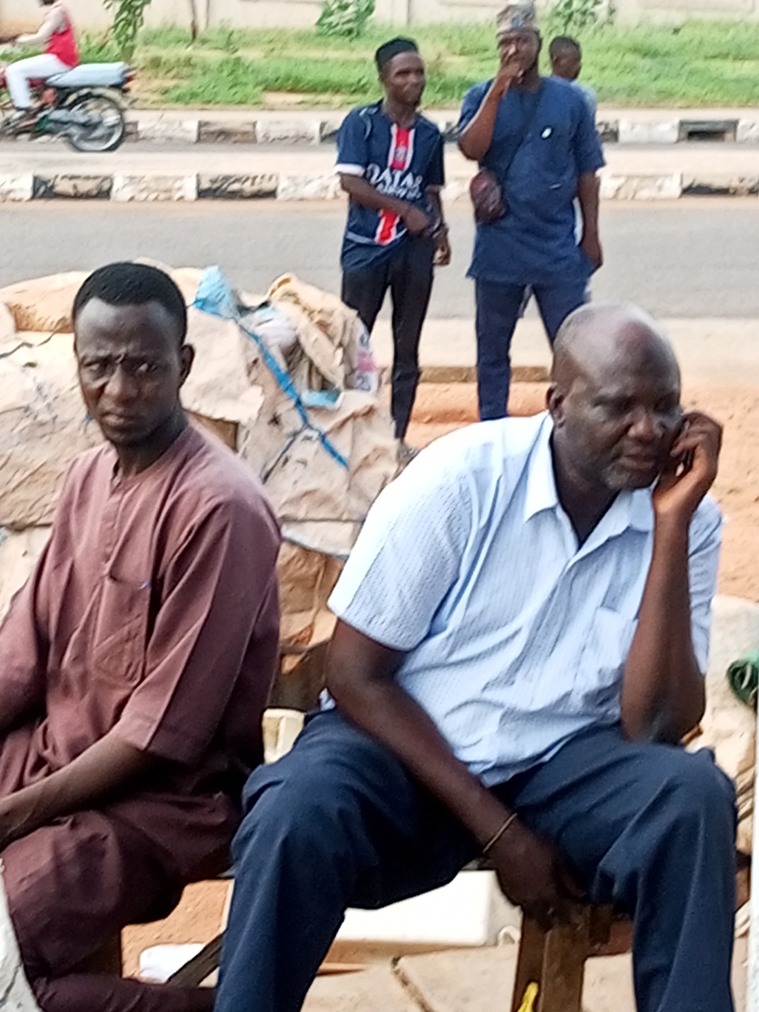 The image depicts an outdoor scene in Gombe, Nigeria. In the foreground, two men are seated. The man on the left wears a maroon traditional tunic. He is looking directly at the camera with a serious expression. The man on the right, with a shaved head, is wearing a light blue collared shirt and dark trousers. He is holding a mobile phone to his ear, engaged in a conversation. His hand rests on his knee, and a thin bracelet is visible on his wrist. Behind them, more individuals are standing. One man in the middle ground wears a blue traditional outfit and a patterned cap, and is holding something to his face as if smoking. To his left, another man wears a blue and white striped t-shirt and dark trousers. In the background, a person is riding a motorcycle. The setting appears to be a roadside area, with bags or goods piled up behind the seated men, suggesting a market or storage area. The ground is dusty, and vegetation is visible in the far background. The lighting suggests it is daytime, possibly late morning or early afternoon.