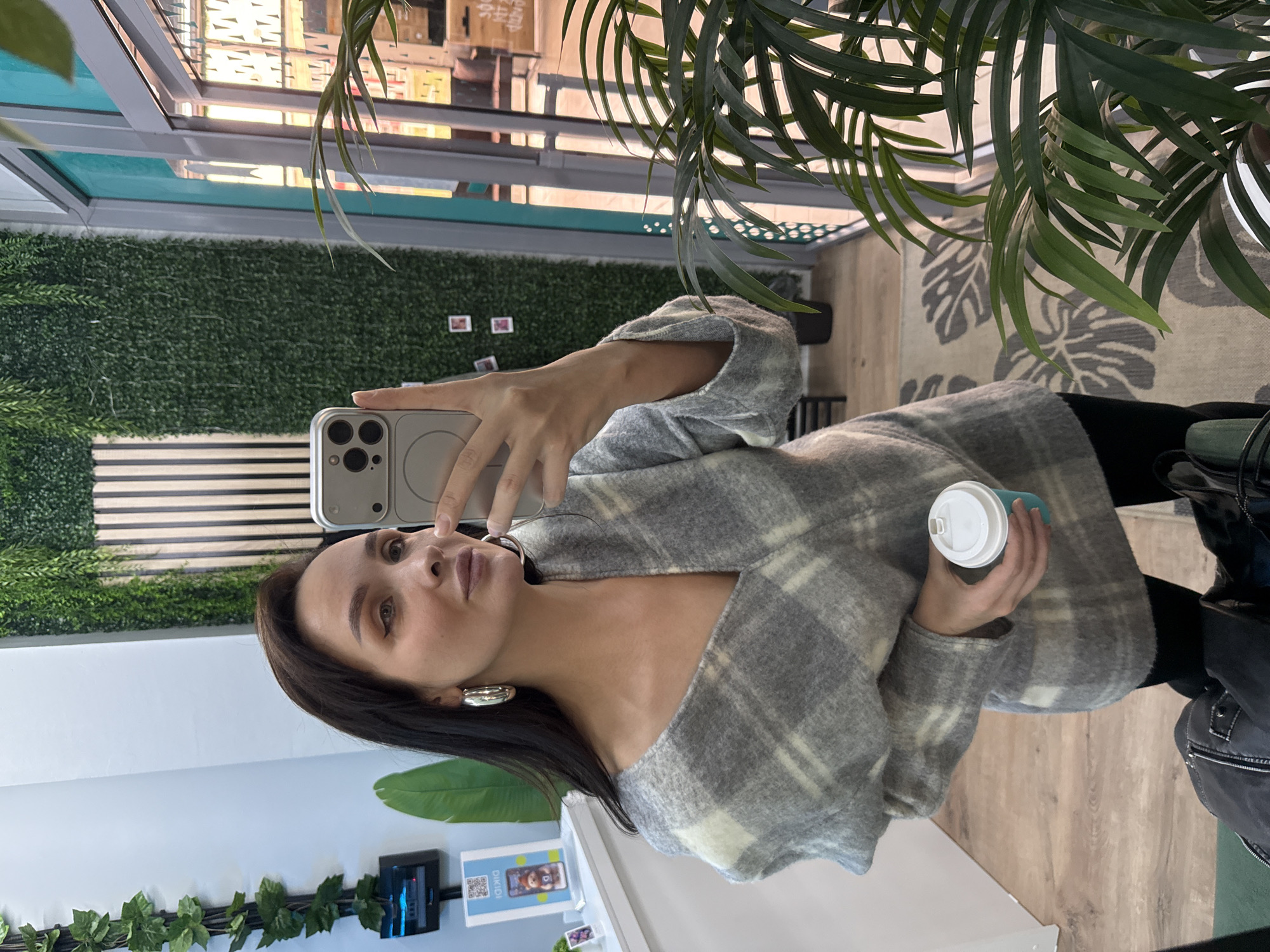 A woman is taking a selfie in what appears to be a cafe or a store. She is holding a phone in one hand and a cup of coffee in the other. She is wearing a gray and white plaid jacket and large silver hoop earrings. Her hair is dark and pulled back. The background shows a wall with green foliage, a window with a view of the outside, and a plant with long green leaves. There are also striped decorations in the background. Based on the presence of the DiKidi app ad, the location could potentially be Chaikovsky, Russia.
