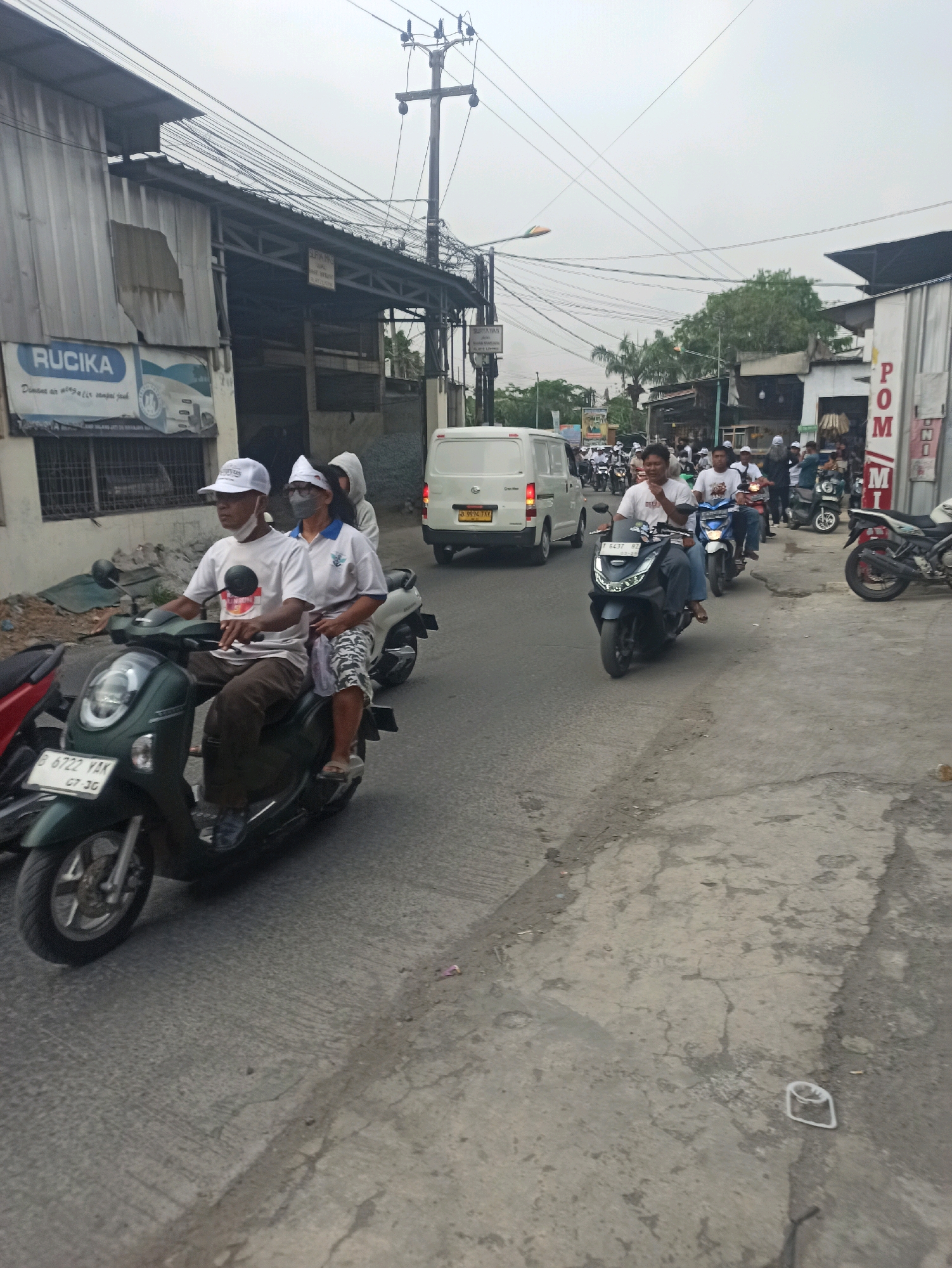 The image depicts a street scene in Wanajaya, Indonesia, characterized by overcast skies and a busy roadway. In the foreground, a dark green scooter carries two individuals. The male rider, wearing a white cap and mask, is dressed in a white t-shirt and brown pants. The female passenger, also wearing a white cap and mask, is dressed in a white polo shirt and patterned shorts. Behind them, a white van is visible, along with numerous other motorcycles and riders navigating the street. The environment includes buildings with corrugated metal siding and a sign that reads "RUCIA". Power lines crisscross the sky, supported by utility poles. The road surface is asphalt with visible cracks and debris. The overall impression is one of everyday urban life in a tropical climate.