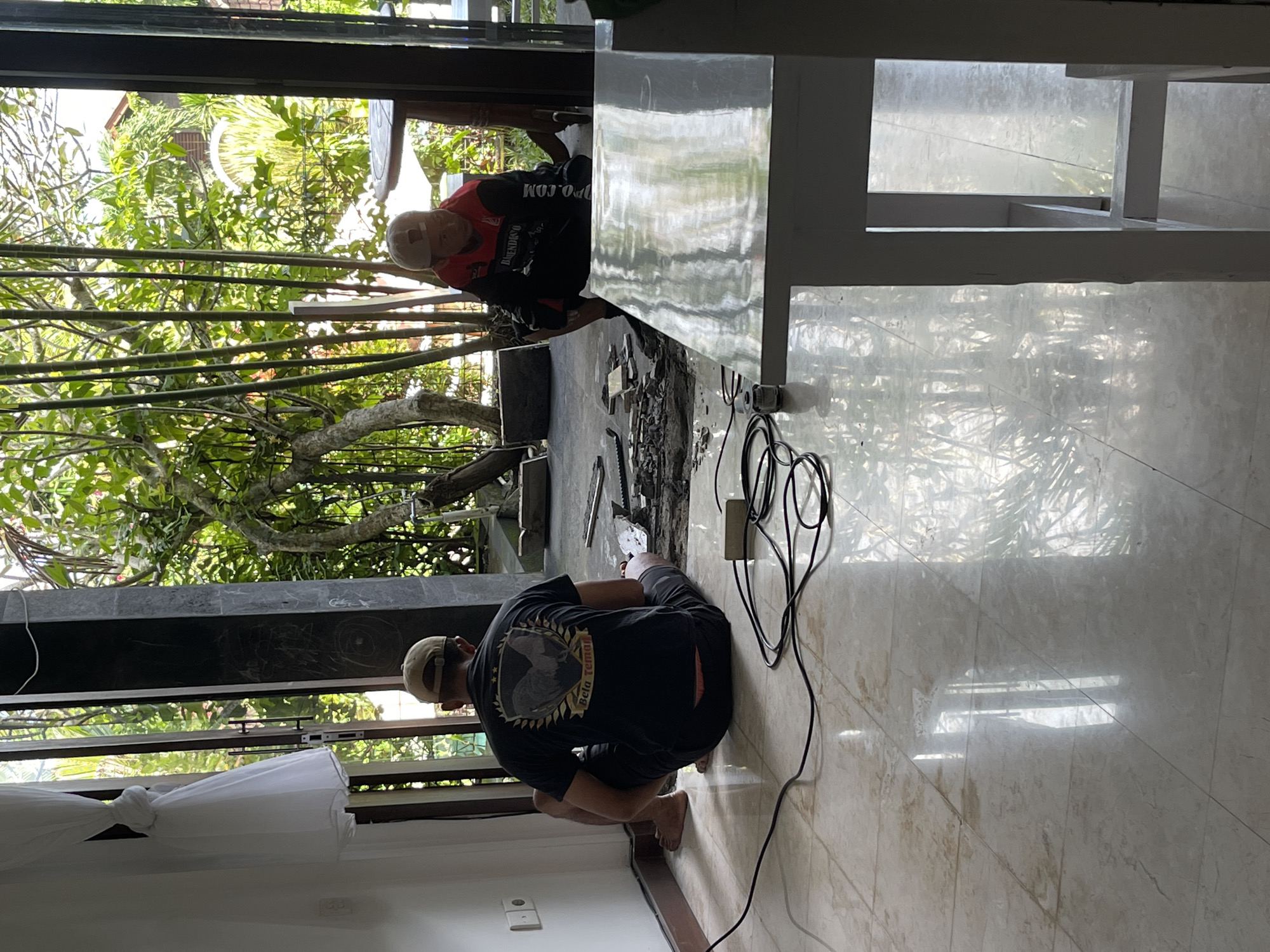 The image shows two men working indoors on a construction or renovation project. One man, wearing a black and red shirt and a light-colored helmet, is positioned near a large window or opening looking out onto a lush, green outdoor area. The other man, wearing a black t-shirt with a graphic design on the back and a light-colored cap, is kneeling on the tiled floor and appears to be operating a tool with an electrical cord. The immediate surroundings include construction debris and the tiled floor reflects light, suggesting a clean, well-lit interior. The exterior through the window reveals dense vegetation.