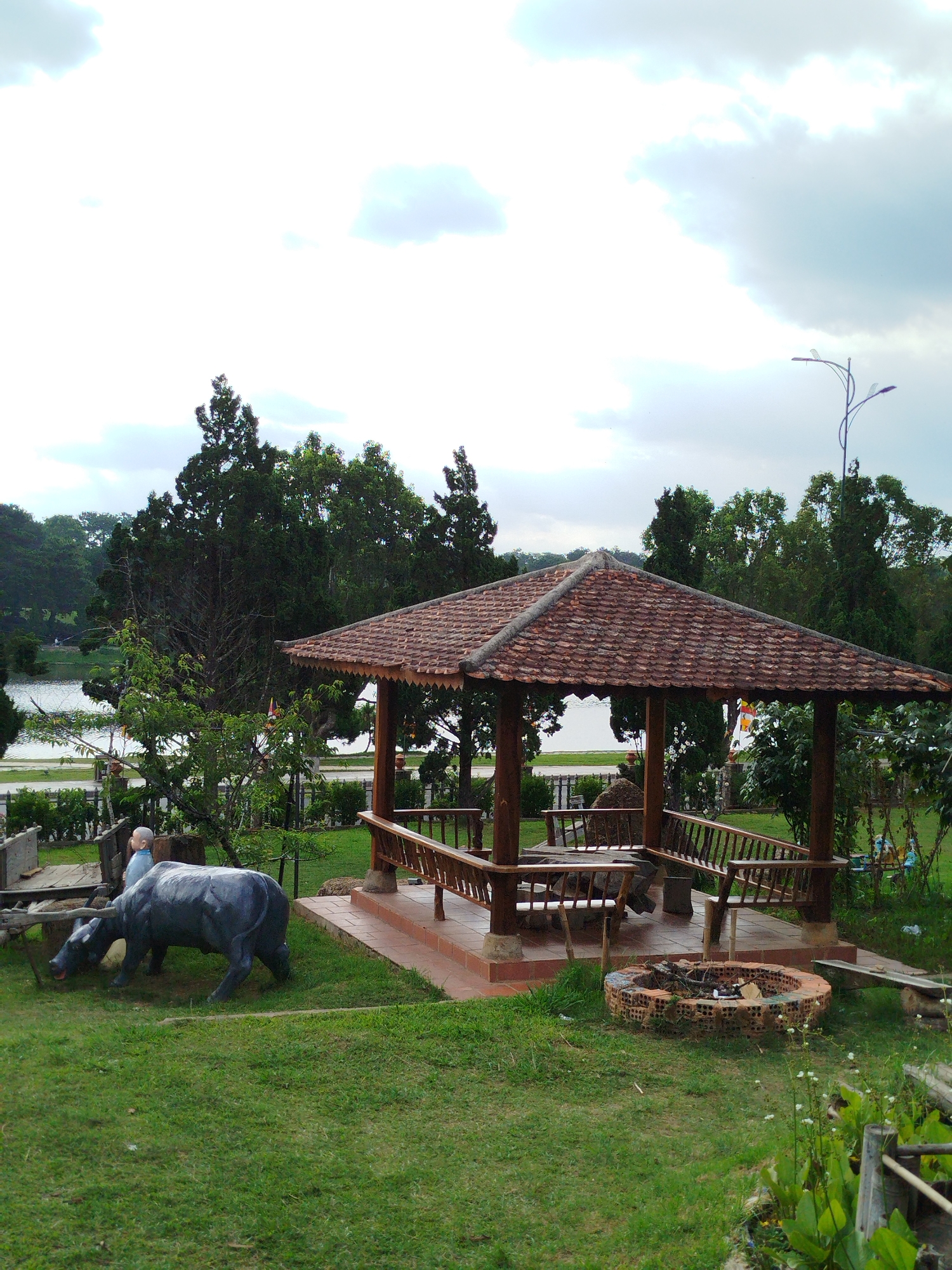 The image shows a park in Da Lat, Vietnam. In the foreground is a green grassy area with a sculpture of a water buffalo and a monk statue. The buffalo sculpture is dark gray and positioned as if it is grazing. The monk statue is next to the buffalo. To the right, there is a fire pit made of brick with some debris inside.
Behind the foreground is a wooden gazebo with a tiled roof, supported by wooden pillars. Inside, there are wooden benches. Someone is sitting on one of the benches.
Beyond the gazebo, there are trees and bushes. In the background, a lake and more greenery can be seen, with a skyline visible in the distance. The sky is overcast with gray clouds. A modern street lamp is visible on the right side of the image.