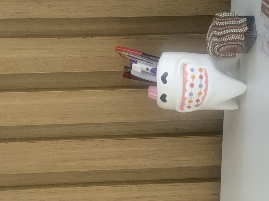 The image displays an indoor scene situated in Lauro de Freitas, Brazil. A white, ceramic, tooth-shaped container, approximately 15 cm in height, stands on a white, flat surface. The container is designed with a cartoonish face, featuring a wide pink mouth with colorful orthodontic braces depicted as multiple circular dots (orange, purple, and pink). Two small, black heart-shaped outlines are present above the mouth, serving as eyes. The container holds multiple writing instruments, including pens and pencils of various colors (e.g., red, white, purple). To the immediate right of the tooth-shaped container, a dark, rectangular object, possibly a stamp or ink pad, is partially visible. It is topped with a textured cover composed of interwoven or beaded strands in shades of brown, pink, and white. This cover features a central circular design with the letters "DR" inscribed. The background consists of vertical light brown wooden paneling with visible grain. No individuals are present in the scene. The environment appears well-lit by ambient indoor light.