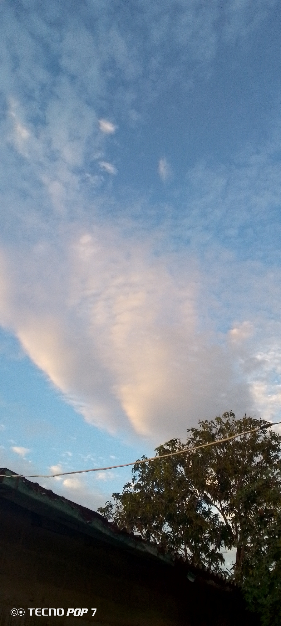 The image depicts a sky with scattered clouds, some tinged with pink from the setting sun. A large, elongated cloud formation stretches across the upper portion of the frame, appearing almost like a celestial wing. In the foreground, the dark silhouette of a rooftop with a corrugated metal surface is visible on the left, with the edge of a tree canopy on the right. A thin wire runs horizontally across the scene, above the rooftop and tree. The sky is a clear blue, transitioning to lighter hues near the horizon. The overall impression is one of a peaceful evening sky over a residential area. The text "TECNO POP 7" is watermarked on the bottom left of the image, indicating the device used for capture. No people or explicit actions are visible.