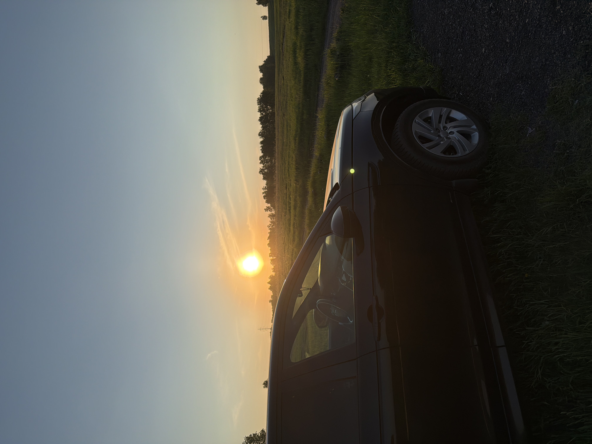 The image depicts an outdoor scene at sunset, featuring a dark-colored SUV positioned on an unpaved track beside expansive green fields. The sky above is clear, transitioning from a soft blue at the zenith to vibrant yellow and orange hues near the horizon, where the sun is visible as a brilliant, intense orb. The low angle of the sun backlights the scene, casting long shadows and creating a bright glare on the vehicle's interior.
A dark-colored SUV, identifiable as a Land Rover Discovery Sport, is parked, with its driver's side profile visible from the front bumper to just past the rear passenger door. The front wheel, equipped with a silver multi-spoke alloy rim and an all-season tire, is in clear view. Sunlight illuminates the interior through the driver's side window, highlighting portions of the steering wheel and dashboard. A distinct, bright green circular reflection or indicator light is visible on the front left fender.
The vehicle rests on a dark, gravel or dirt track that extends out of frame to the right. To the immediate left of the vehicle and along the edge of the track, tall, green grass flourishes. Beyond the track, verdant fields stretch into the distance, intersected by a faint dirt path running horizontally across the mid-ground.
Along the distant horizon, a dense line of deciduous trees forms a dark silhouette against the bright sky, particularly on the left side. A solitary tree stands on the far right horizon. A slender, vertical structure, possibly a utility pole or transmission tower, is faintly visible above the treeline in the far-left background. No individuals are present in the scene, and no specific actions or interactions are occurring.
The environment depicted is characteristic of rural or semi-rural landscapes, indicative of agricultural fields and unpaved roads, consistent with areas found on the periphery or outskirts of Krasnoyarsk, Russia, rather than within its densely built-up urban center. Specific street names or identifiable architectural landmarks of Krasnoyarsk are not discernible from the visual content.