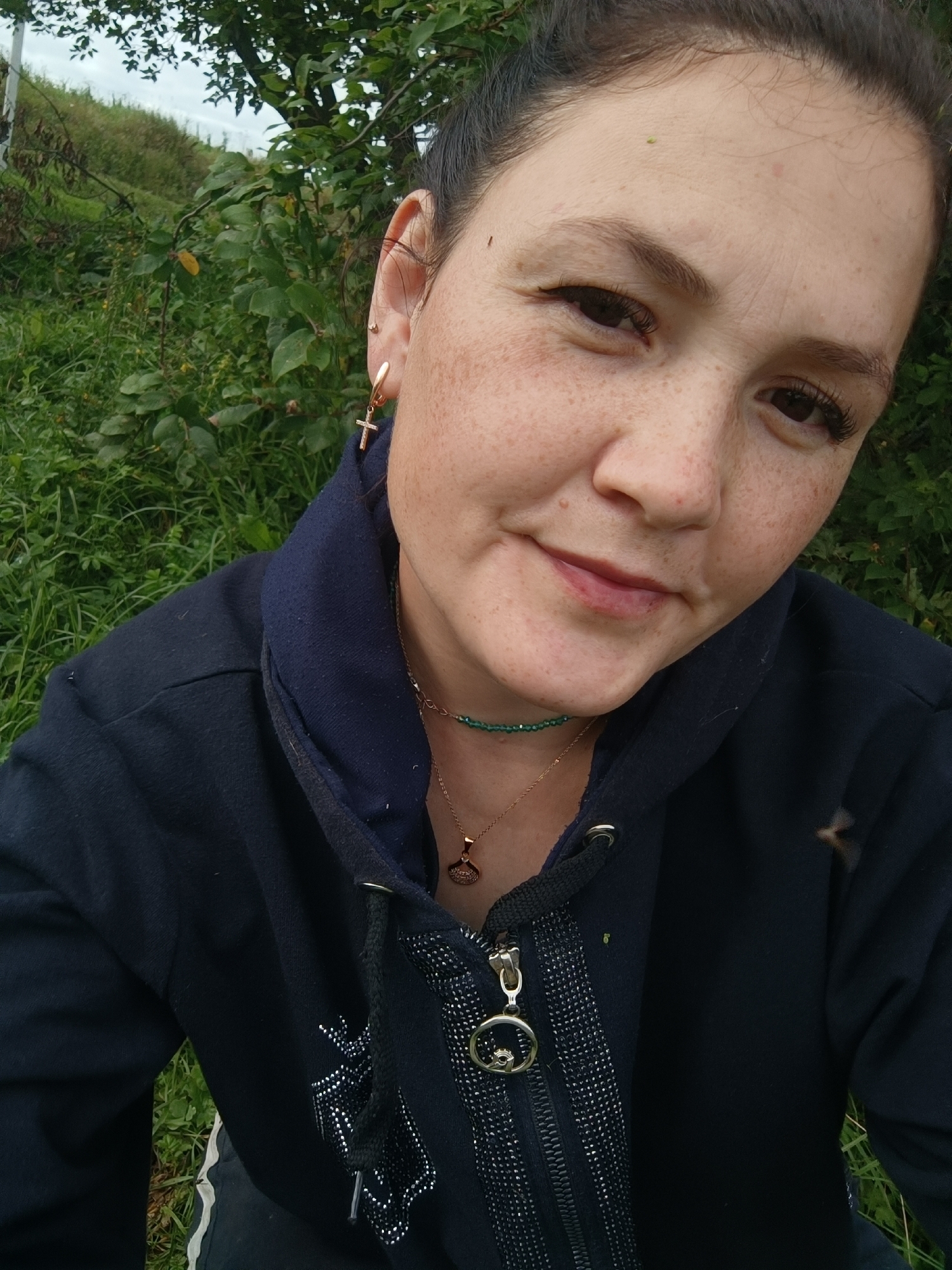 The image is a close-up, portrait-style photograph of a woman outdoors. She is looking towards the camera with a slight smile. The background consists of green foliage and trees, suggesting a natural, outdoor setting. The woman is wearing a dark blue hooded jacket, a necklace with green beads, and a gold pendant. She has a cross-shaped earring in her left ear. Her face is visible, with freckles and dark hair pulled back. The lighting appears to be natural daylight. No specific landmarks or urban features are visible to indicate a location in the city of Karsha, Russia.