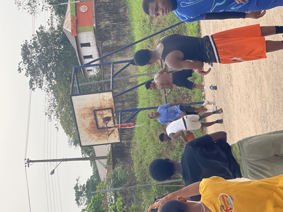 An outdoor scene in Umunekwu, Nigeria, depicts a group of approximately eight young men engaged in a game of basketball on a dirt court under an overcast sky. The focal point is a visibly aged basketball hoop with a rusty backboard, a blue metal frame, and a partially torn red and white net.
In the mid-ground, three players are active on the court. One player, wearing a white tank top and light-colored shorts, dribbles an orange basketball. To his left, another player in a blue t-shirt and black shorts stands with a wet shirt, observing. To the right, a shirtless player in black shorts stands, looking towards the dribbler.
In the foreground, from left to right, four individuals are observing the game. One person, largely out of frame, wears a yellow t-shirt and holds a phone up, seemingly filming or photographing the game. Next to them, a person in a black t-shirt and light green shorts stands with their back to the camera. Further right, another individual with their back to the camera wears a black tank top and orange shorts. On the far right, a player in a blue patterned long-sleeve top looks towards the court action.
The environment includes overgrown green grass surrounding the court. In the background, a concrete wall separates the court area from a small, white building with a red and green roof. A sign on the building is partially visible, reading "Daily Bread." Above the building, a utility pole with electrical wires and a single street light is prominent against the sky. Lush green trees and foliage are visible beyond the structures.