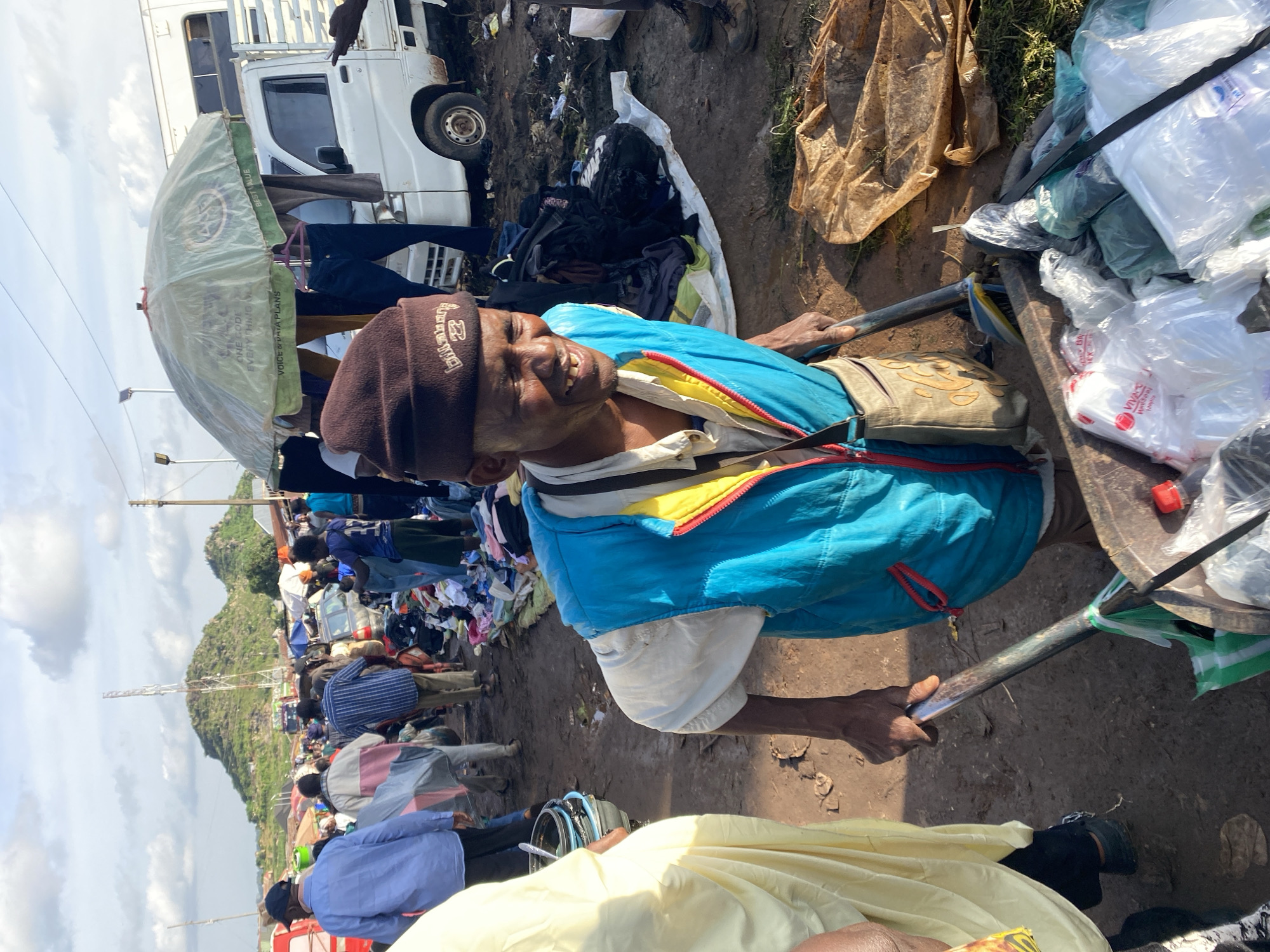 The image depicts a bustling outdoor market scene in Gura Top, Nigeria, under a partially cloudy sky. The foreground is dominated by a man of African descent, wearing a brown beanie and a blue and yellow vest over a white shirt. He is smiling and pushing a cart filled with plastic bags and other items. His right hand grips the handle of the cart, which is also laden with a broom and what appears to be a shovel. Behind him, a woman in a yellow garment is partially visible on the left.
The background reveals a crowded market with numerous people browsing various stalls and displays of goods, primarily clothing, laid out on the ground. Several vehicles, including a white truck and red trucks, are present in the market. In the distance, rolling hills with rocky outcrops and a prominent telecommunications tower can be seen. The ground is predominantly dirt and mud, suggesting recent rain or the general conditions of the market. An umbrella, advertising "ONE CODE EVERY THING VODACOM," is also visible, providing shade over some of the stalls. The overall impression is one of lively commerce and activity within a semi-urban environment.
