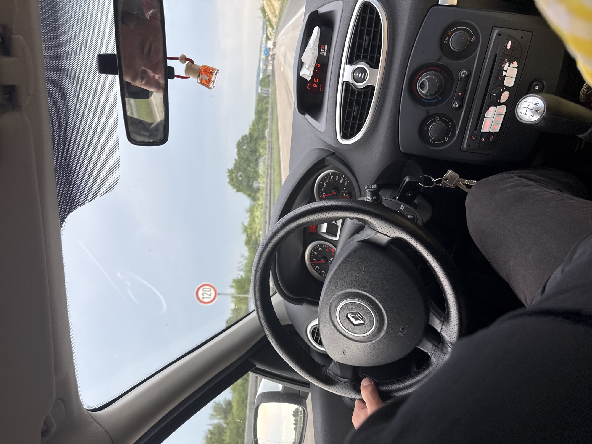 The image shows the interior of a car driving on a highway. A person's hands are visible on the steering wheel, indicating they are the driver. The dashboard displays include a speedometer, radio, and air conditioning controls. A speed limit sign of 120 is visible outside the windshield. In the rearview mirror, the face of another person is reflected. The scene appears to be a sunny day, with trees and other cars visible on the highway. The location is identified as Linden, Germany.