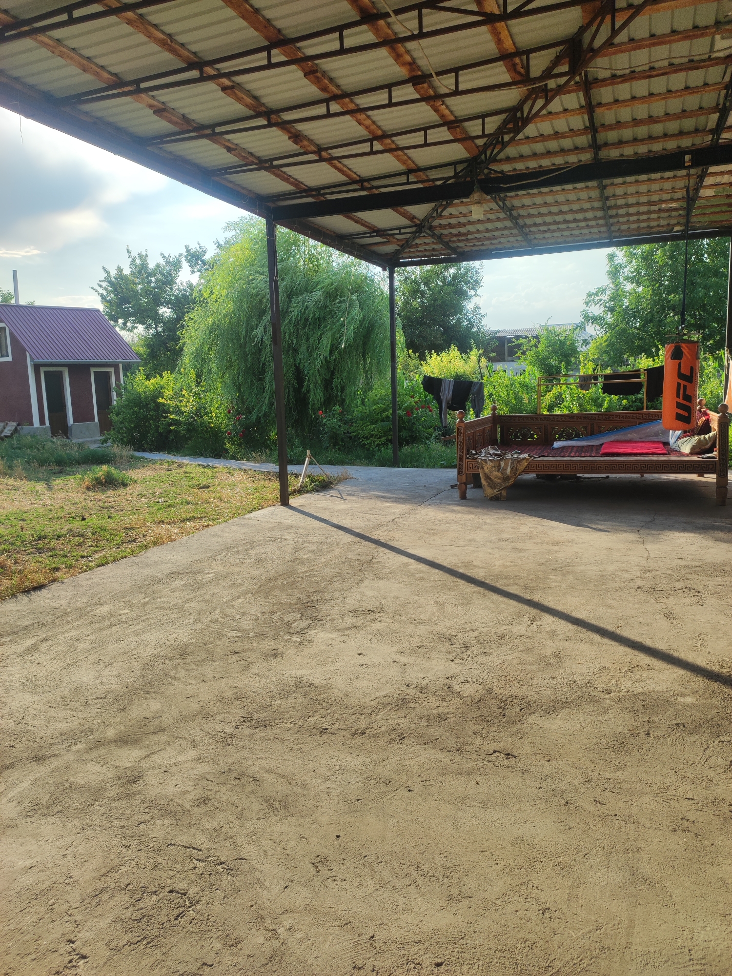An outdoor scene under a metal and corrugated roof structure, possibly a carport or patio. The ground is concrete, with a visible shadow from the roof and a support post. To the left, a small, maroon building with a dark roof stands amidst dry grass and some debris. In the background, lush green trees, including a large weeping willow, are visible. A wooden daybed or bench, adorned with decorative carvings and covered with textiles and cushions, is positioned under the shelter. An orange UFC punching bag hangs from the roof structure above the daybed. There are no people visible in the image. The setting appears to be rural or semi-rural, possibly in the city of Mingchinar (Blagoveshchensky), Uzbekistan.