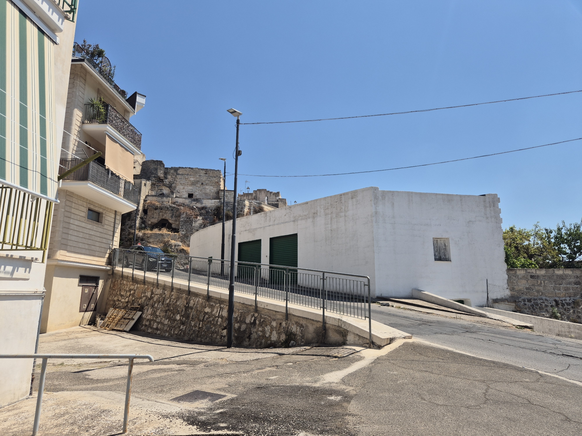 The image depicts a street scene in Laterza, Italy. In the foreground, a paved road curves to the right, with cracked asphalt and scattered debris. To the left, a building with cream-colored walls and a green and white striped awning is visible. A metal railing runs along the edge of the sidewalk.

Further into the scene, a stone retaining wall supports a sloping area behind a metal fence. Above this, a modern white building with two large green garage doors is situated. The building has a small, shuttered window.

In the background, the ruins of an ancient structure are visible on a rocky hillside, suggesting a historical context for the town. The sky is a clear, bright blue. A black car is parked on the street in front of the white building. Two streetlights are present, one taller than the other. Power lines are strung across the sky. The environment appears to be urban with elements of historical architecture and natural landscape.