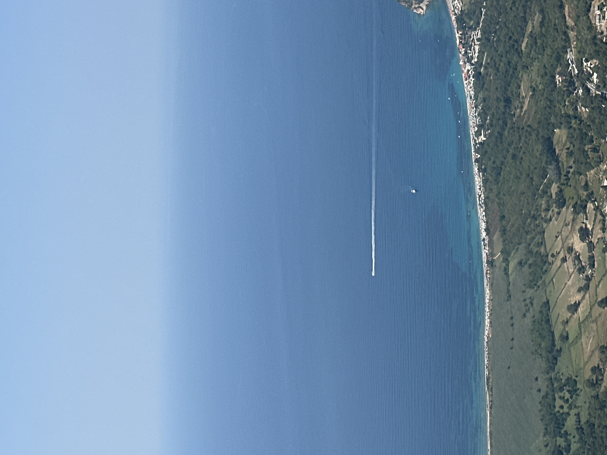 This is a high-angle aerial shot of a coastline in Gluhi Do, Montenegro, on a clear day. The vast expanse of the Adriatic Sea dominates the upper portion of the image, transitioning from a deep blue to a lighter, turquoise hue near the shore. A white boat is visible in the distance, leaving a faint wake. Closer to the shore, several smaller boats are scattered. The coastline itself features a long, sandy beach lined with numerous beach umbrellas and a small town or village with buildings nestled among green vegetation. The foreground shows a verdant landscape with patchwork fields and trees, indicative of the hilly terrain surrounding the coastal area.
