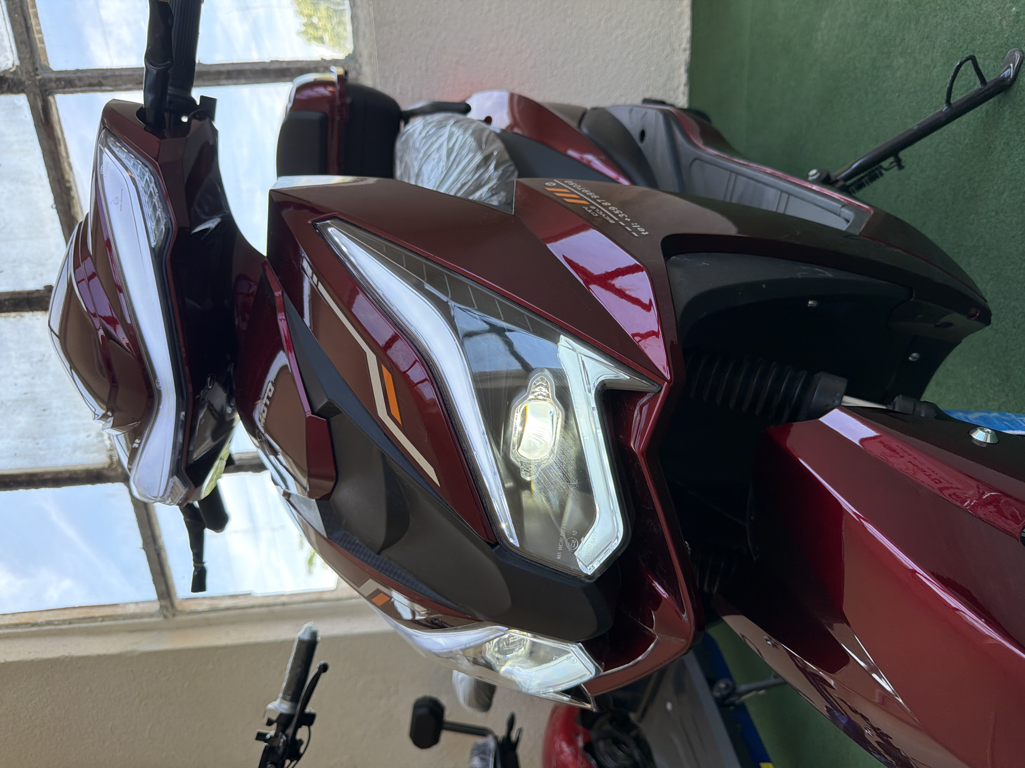 A close-up, low-angle shot captures a dark red electric scooter parked indoors. The scooter's front, including its sharp, modern headlights and handlebars, dominates the frame. The headlights are illuminated, casting a bright glow. A portion of a plastic-wrapped seat is visible behind the front of the scooter, and a black storage box is mounted above it. To the left, another scooter, primarily red, is partially visible, with its seat and handlebars showing. The background reveals a large window with a paneled, possibly industrial-style frame, revealing a bright, overcast sky. The floor is covered with a green mat. The wall to the left of the window is a light beige color. A phone number and the words "CITY BICYCLE" are visible on the scooter's side.