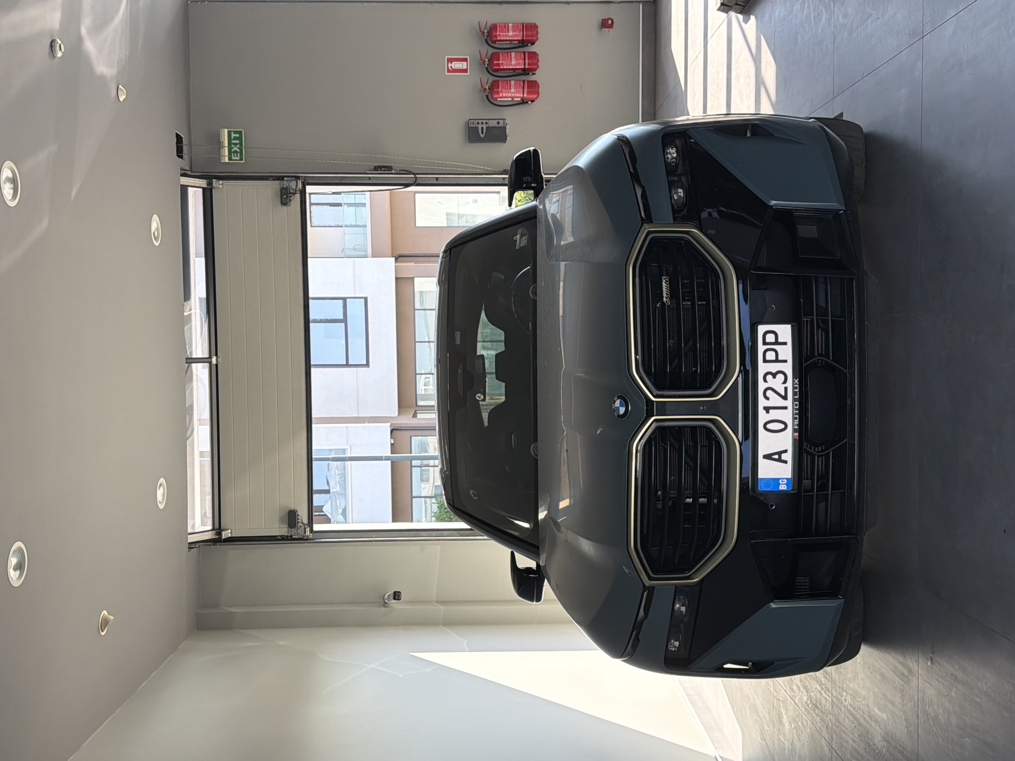 A dark gray BMW SUV is parked indoors in what appears to be a showroom or garage. The vehicle faces the camera, with its headlights on. The license plate is visible, reading "BG A 0123 PP". Behind the car, a large, partially open garage door reveals modern buildings with balconies and windows. To the right of the car, on a gray wall, there are two red fire extinguishers and a gray box mounted on the wall. An illuminated green "EXIT" sign is visible on the wall above and to the right of the fire extinguishers. On the ceiling, there are recessed lights and a small security camera. The floor is tiled with large gray tiles. The scene is brightly lit, with sunlight casting shadows on the floor from the open garage door.