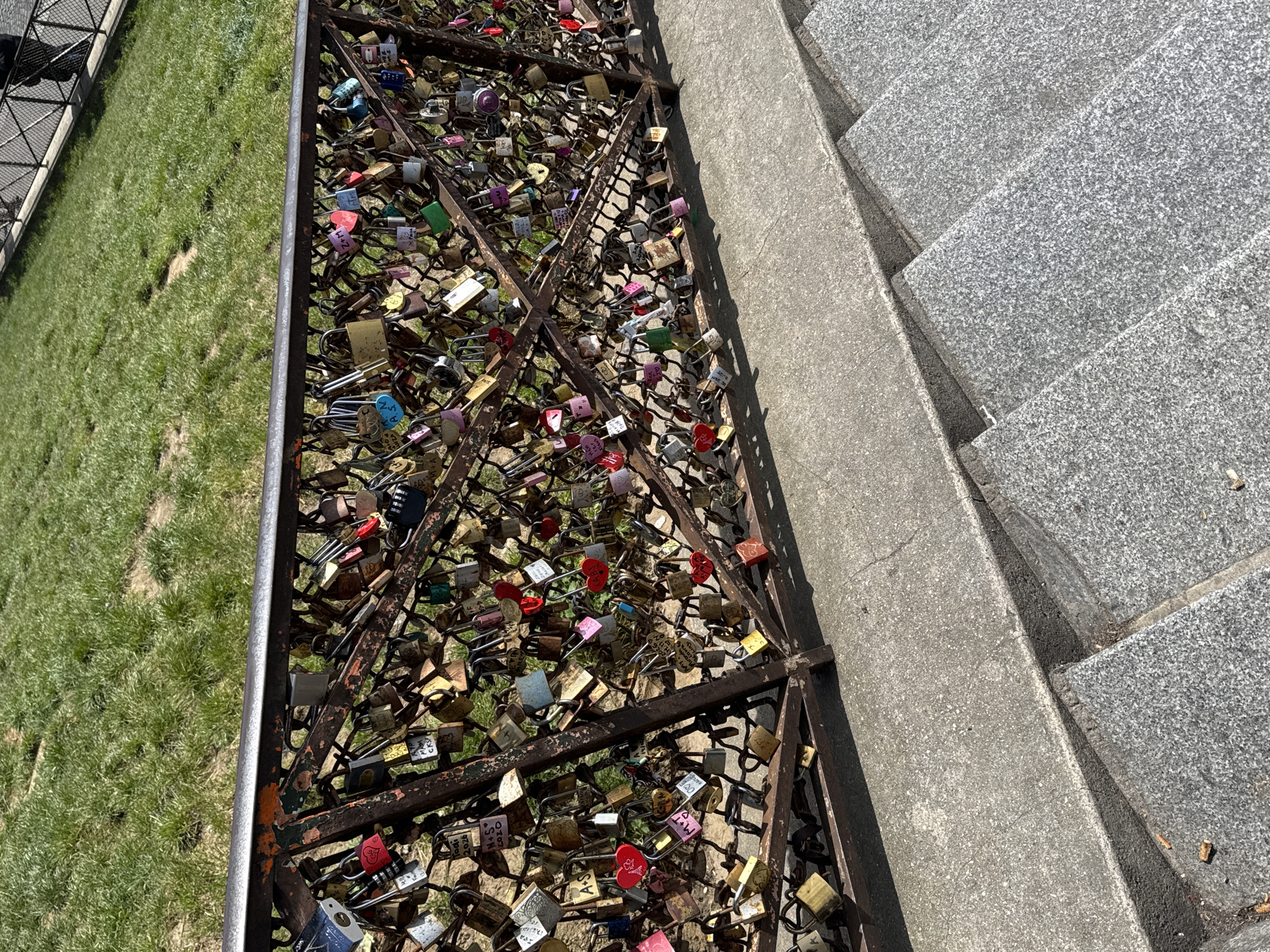 The image depicts a section of the "love lock" bridge in Paris, France, viewed from an elevated position, likely the adjacent steps leading up to the bridge. The foreground contains multiple rows of gray, granite steps leading to a weathered, metal railing filled to capacity with hundreds of padlocks. The padlocks vary in size, shape, color, and material (brass, steel, etc.) and appear to have been attached by couples as a sign of their love. There is a section of grass to the left of the rail. The image suggests a sunny day. No people are visible.