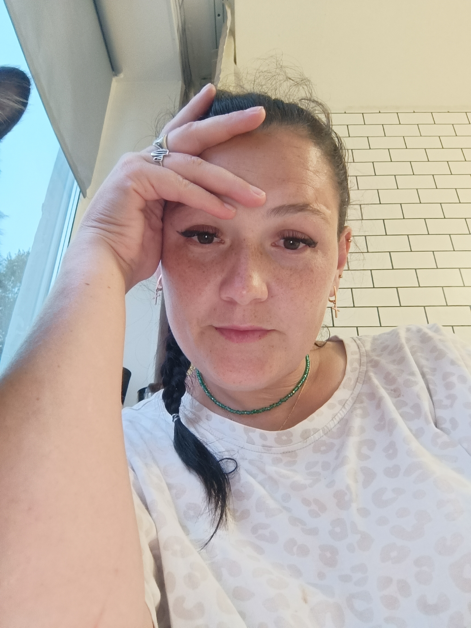 The image shows a close-up, low-angle selfie of a woman with dark hair pulled back and braided. She is resting her forehead on her hand, with her fingers splayed across her brow. She is wearing a white t-shirt with a subtle leopard print pattern and a green beaded necklace. She has noticeable freckles and is wearing mascara and winged eyeliner. She is also wearing a silver ring with an abstract design on her right index finger and small hoop earrings. The background features a tiled white wall and a window with a view of trees. The location is specified as being in the city of Chaikovsky, Russia.