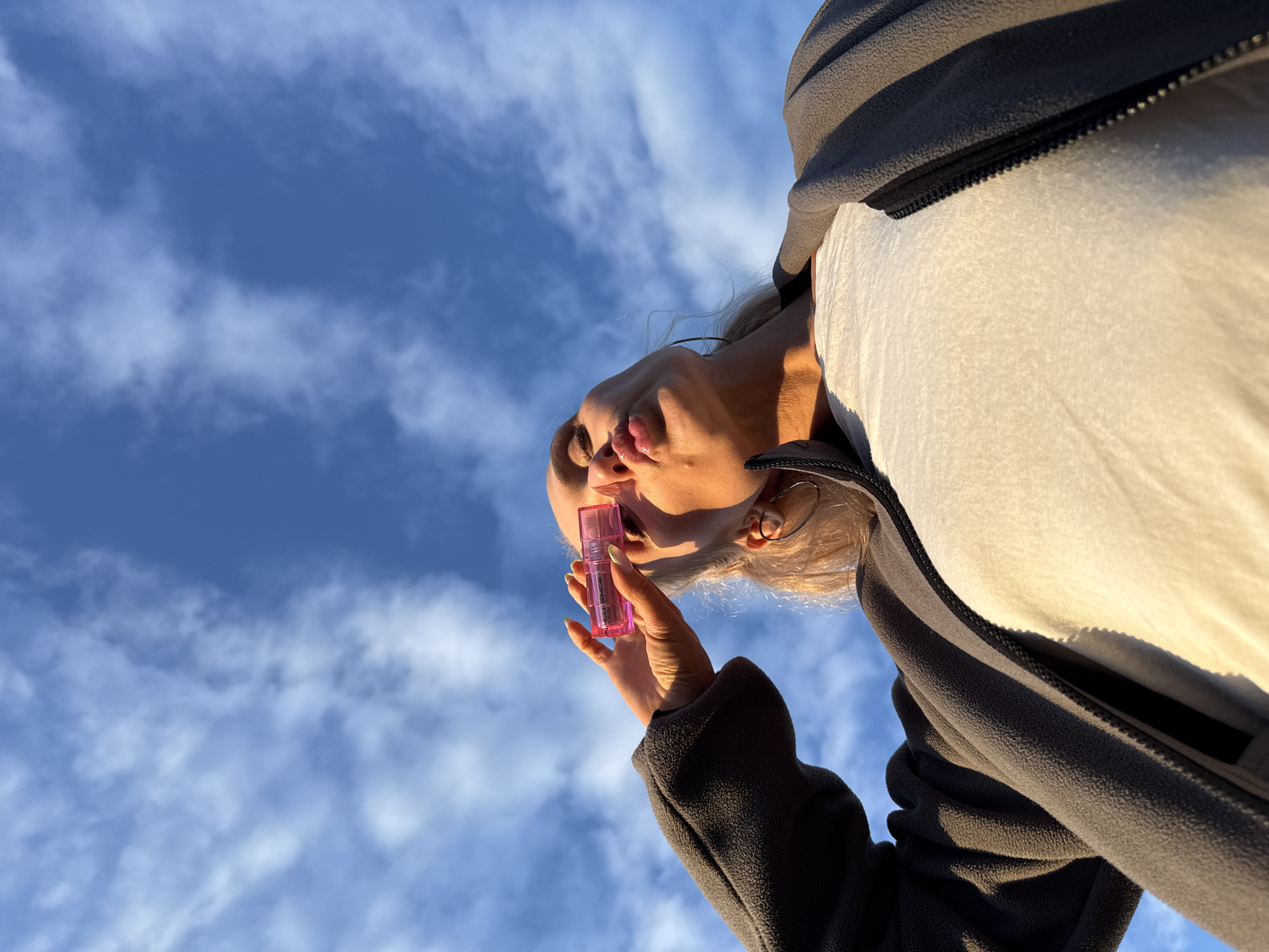 The image is a low-angle, upward-facing shot featuring a person against a clear blue sky with scattered white clouds. The person, visible from the upper chest, has light-colored hair and is wearing a dark gray fleece jacket with a visible black zipper over a light-colored top. In their right hand, they hold a small, rectangular, translucent pink cosmetic product, possibly a lip gloss or similar item, with the word "INFLUENCE" visibly printed on it. The product is held close to the right side of their face, near the eye and nose. The person's lips are pursed in a "kiss" pose, and their eyes are partially closed, looking downward towards the held object. A large, thin, silver-colored hoop earring is visible on their left ear. The lighting suggests direct sunlight, creating strong highlights and shadows on the person's face and jacket. The nails on the visible hand are painted, some with a light base and a distinct bright yellow or green tip. There is no visual information within the image to indicate its specific geographical location in the city of Chaikovsky, Russia, or anywhere else.