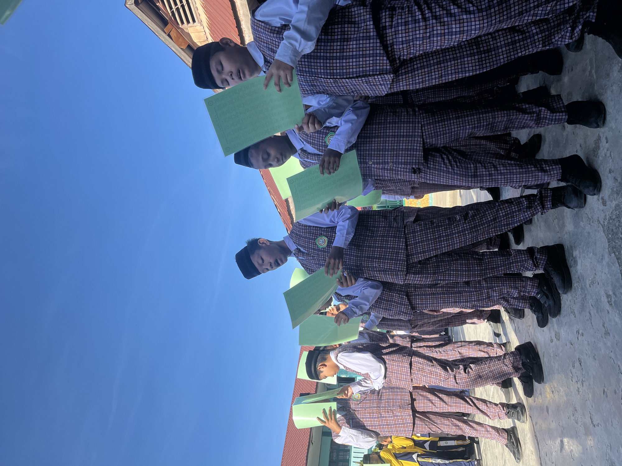A group of young boys, dressed in plaid vests, white shirts, and black peci caps, stand in a line outdoors, holding green papers. The boy in the foreground on the right is looking up and singing with his mouth open. The boys are standing on a paved surface in front of a building with an orange roof. The sky is clear and blue. In the background, to the left, other children in yellow and blue outfits can be seen. The scene appears to be taking place at a school in Lhokseumawe, Indonesia.