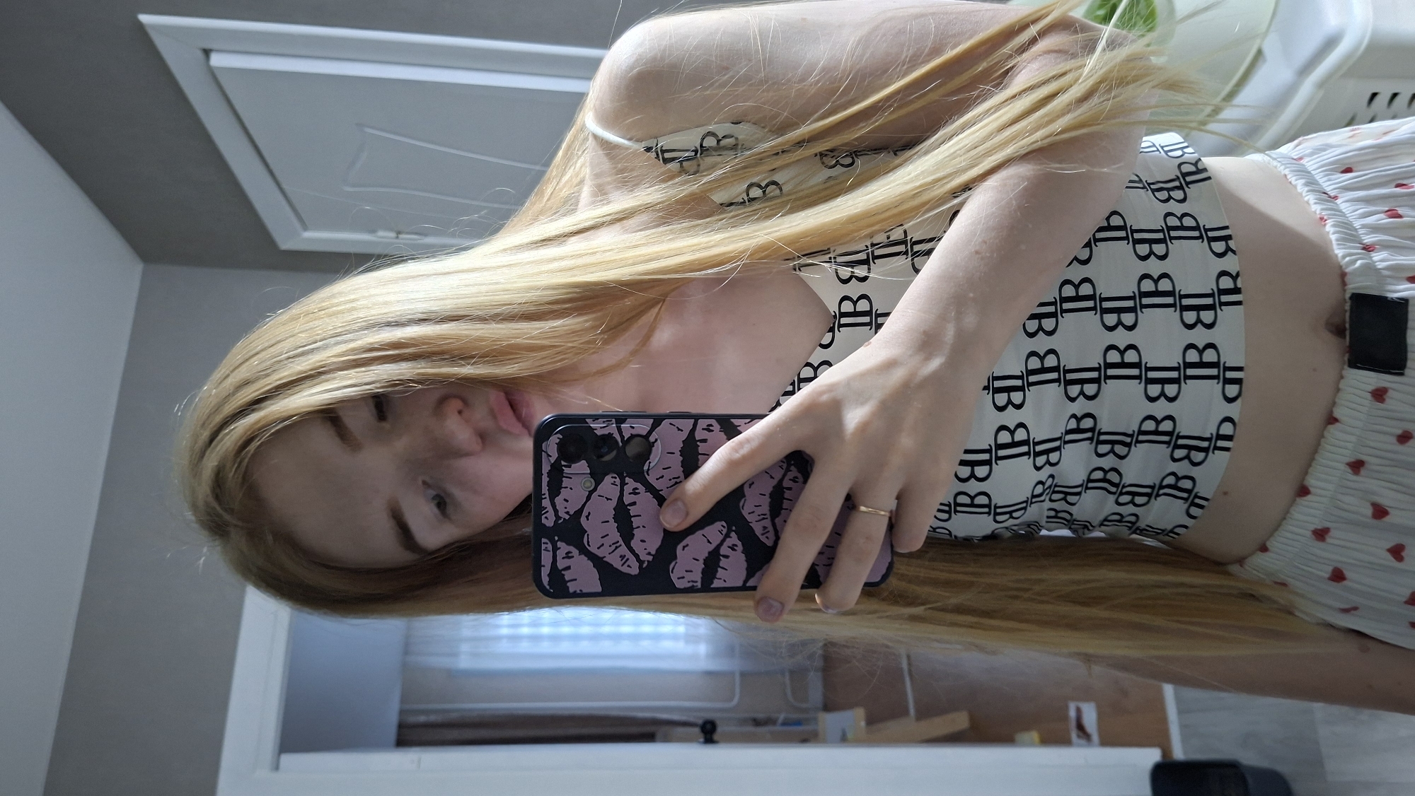 Here is a detailed description of the image:
**People:**
* A fair-skinned person with long, blond hair is the main subject.
**Objects:**
* A mobile phone is being held by the person. The phone case has a design of dark-colored lips.
* The person is wearing a white top with a repeating "B" pattern, and bottoms with a red heart pattern.
* A gold-colored ring is visible on one of the person's fingers.
* A closed door is visible in the background.
**Environment:**
* The scene appears to be indoors, possibly in a bedroom or bathroom.
* The location is the city of Chaikovsky, Russia.
**Actions:**
* The person is holding the phone and taking a selfie or looking in a mirror.
* The person is tilting their head.
