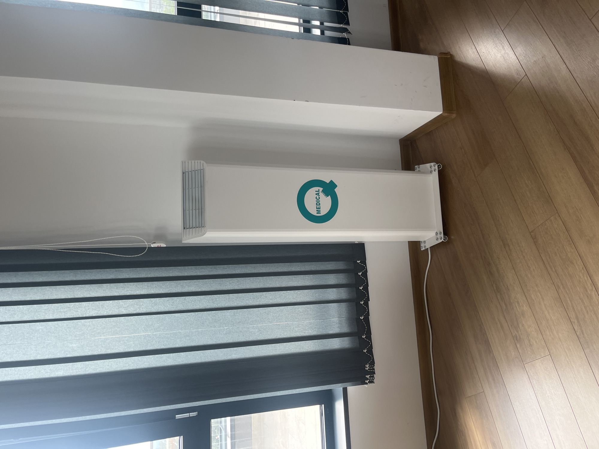 The image shows a room interior with a white radiator positioned under a window with gray horizontal blinds. The radiator has a "MEDICAL" logo on it. The room has light brown wooden flooring. The scene is indoors, likely a room in a building in Plovdiv, Bulgaria, though there are no specific external landmarks visible through the window to confirm the exact location. There are no people visible in the shot.