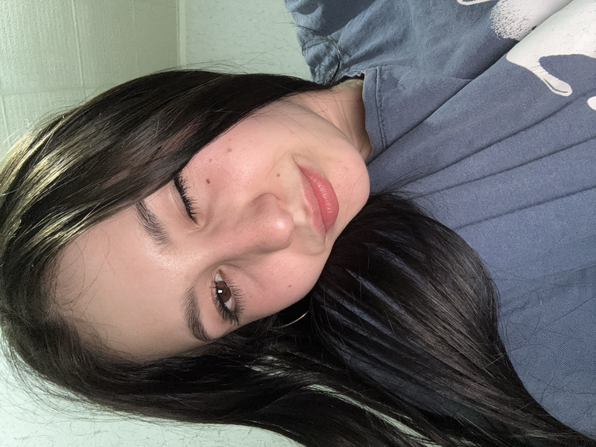 A portrait of a young woman with long, dark hair, wearing a blue t-shirt with a white design. Her head is tilted slightly to the side, and she is looking at the camera with a slight smile. Her skin has a few small moles. She is wearing a small hoop earring in her right ear. The background is a light, solid color. There is no evidence of the location being the city of Фоки, Russia.