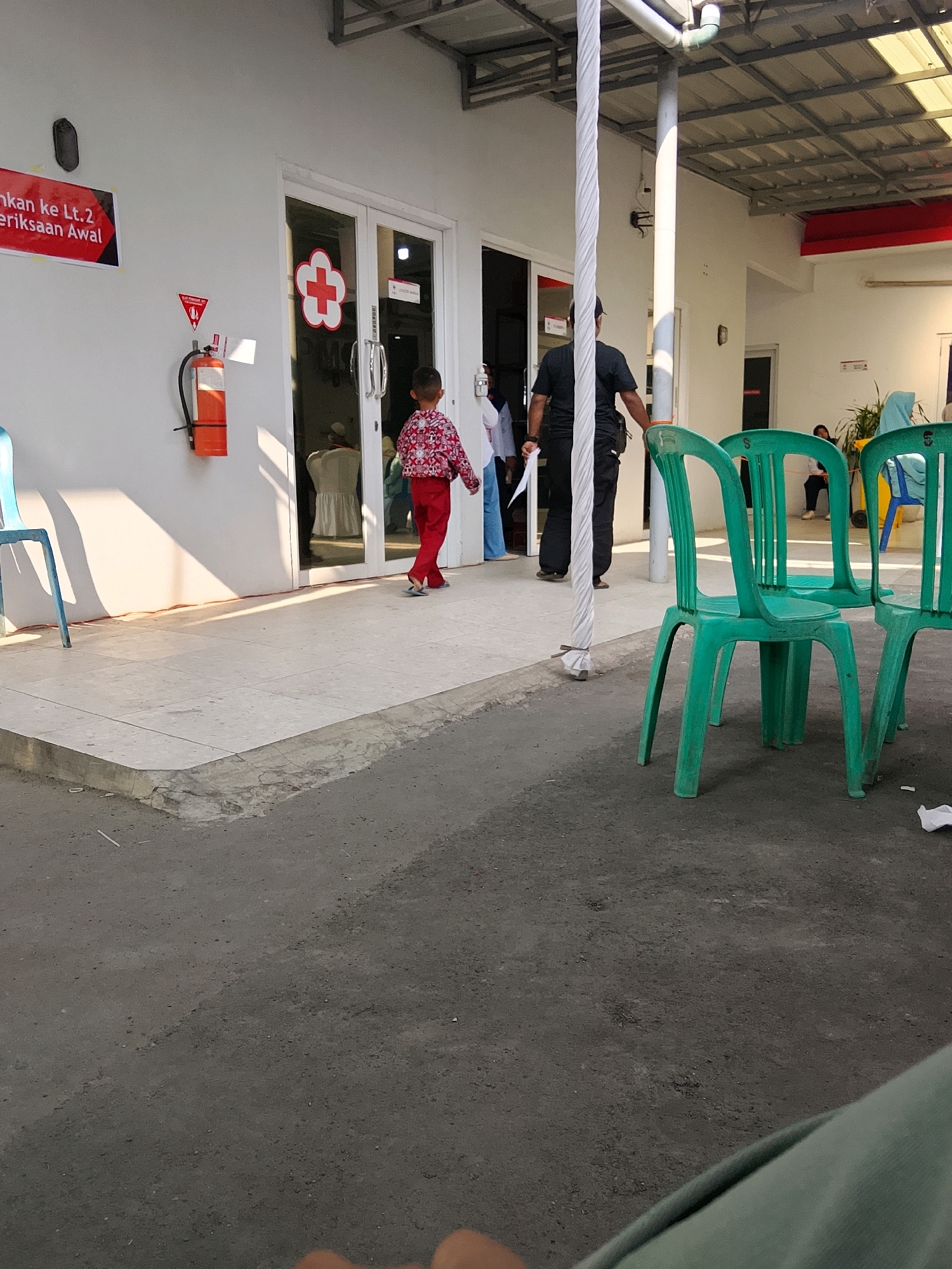 The image captures an outdoor scene in Cirebon, Indonesia, likely in front of a medical facility, given the prominent red cross symbol on the building's glass doors and the presence of a fire extinguisher. In the foreground, green plastic chairs are arranged, some of which are occupied by people. A young child in a patterned red and white shirt and red pants is walking away from the viewer towards the entrance. Behind the child, a man in a black shirt and dark pants is walking into the building. Sunlight casts distinct shadows on the tiled walkway and the white wall of the building. Other individuals, including a woman in a blue headscarf and another in a light-colored hijab, are visible seated or standing in the background, suggesting a waiting area. A red and black sign on the wall reads "nkan ke Lt.2 Pemeriksaan Awal," which translates to "Go to the 2nd Floor Initial Examination." The environment appears to be a public space, possibly a clinic or hospital reception area, with a paved ground and a covered walkway.