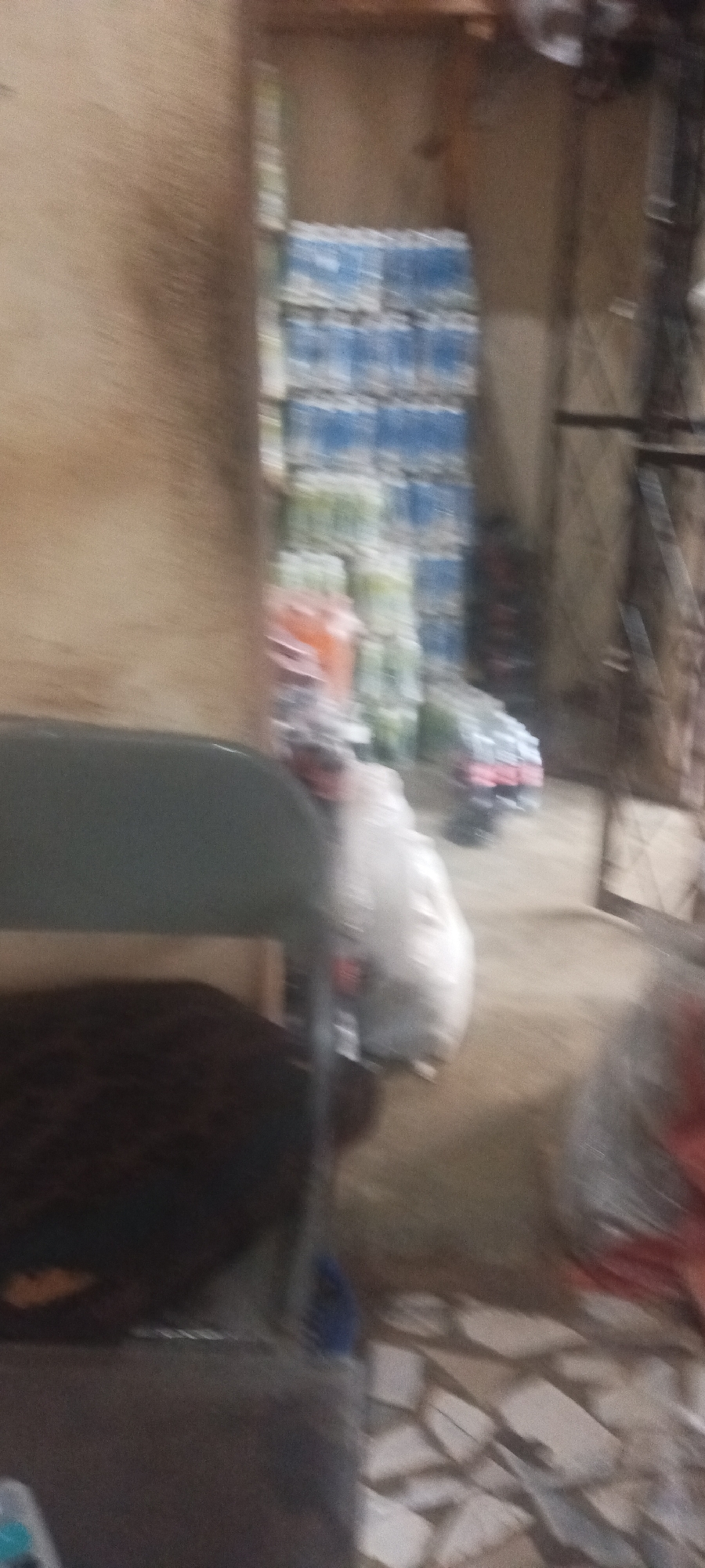 The image, taken in Mangu Town, Nigeria, depicts an indoor storage area. Stacks of bottled water are visible in the background. There are also bags and other objects partially obscured by a gray chair and lack of focus. The floor appears to be tiled with irregular pieces. The image suffers from motion blur.