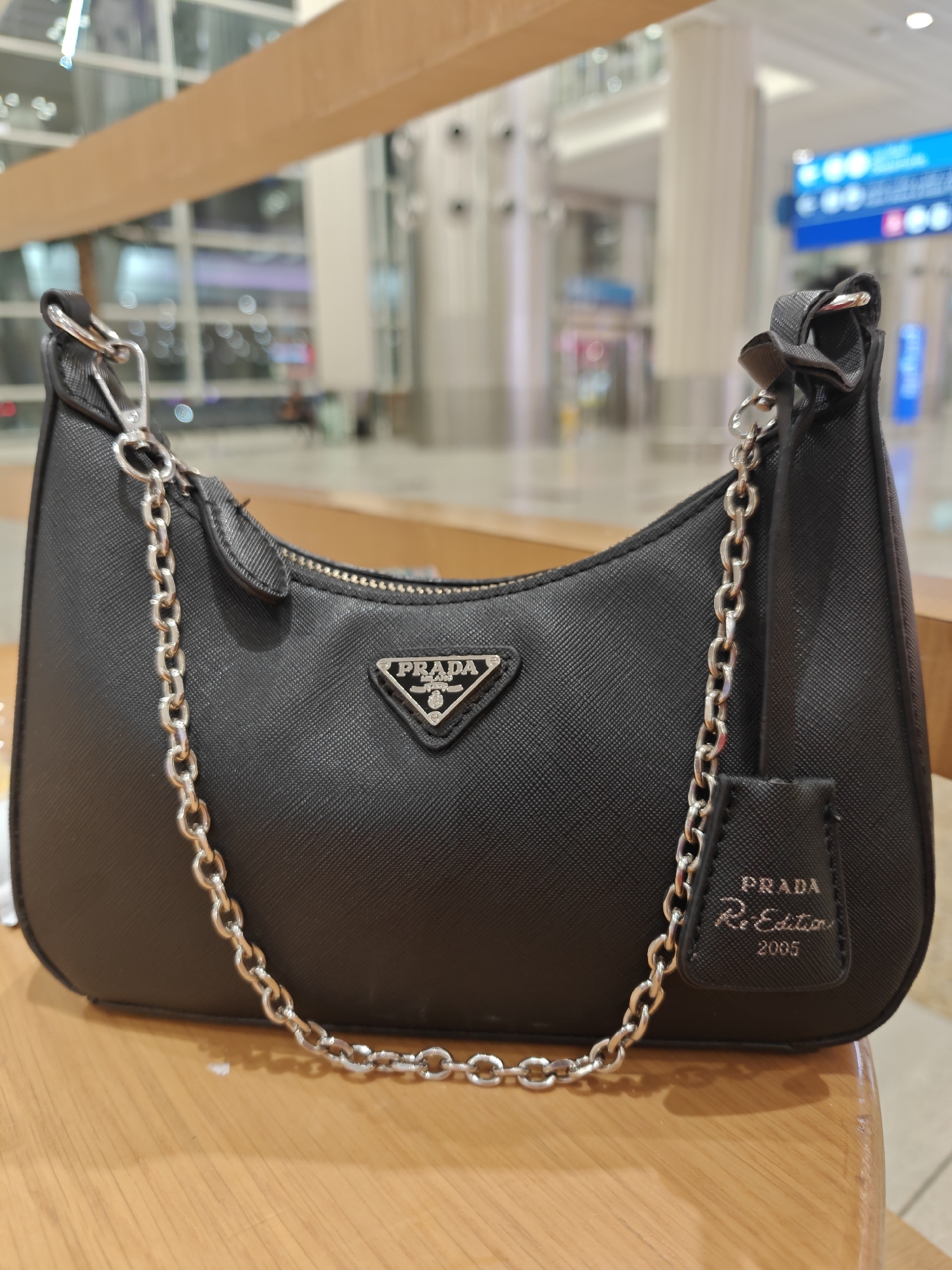 A black Prada Re-Edition 2005 handbag is displayed on a wooden surface. The handbag features a silver-tone chain strap and a silver Prada triangle logo. The background appears to be an indoor airport terminal with blurred figures and signage, suggesting the location is Dubai International Airport, United Arab Emirates.