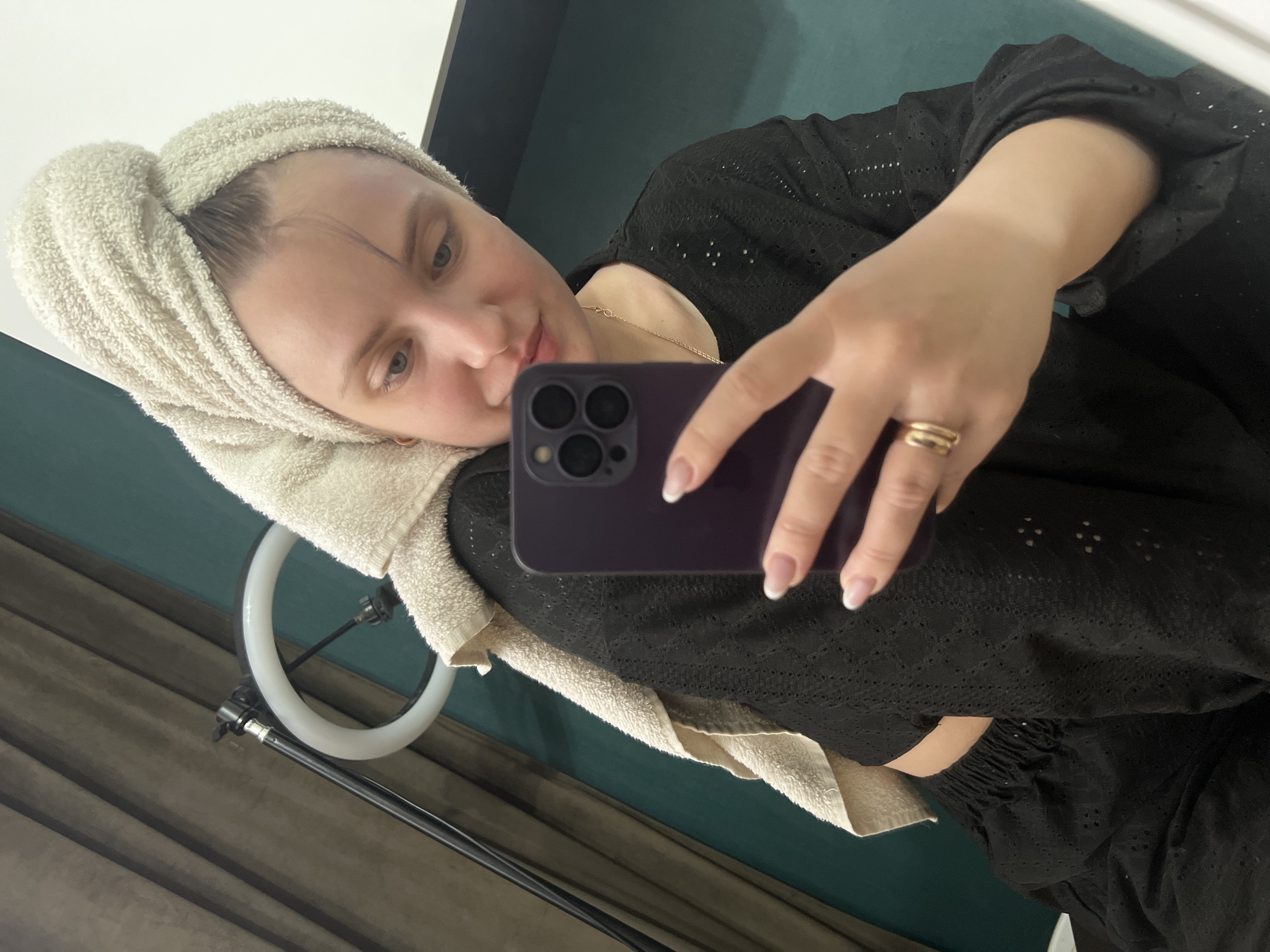 A woman is taking a selfie in what appears to be an indoor setting. She has a towel wrapped around her head and another draped around her shoulders. She is holding a purple smartphone, possibly an iPhone, in her right hand to capture the photo. Her nails are manicured with white tips, and she is wearing at least one ring on her finger. The woman is wearing a black top with a perforated design. A ring light is visible in the lower left of the frame. The background includes a wall with what seems to be a dark teal color above a lighter shade of gray. There is no verifiable evidence to confirm the location as Марковский, Russia.