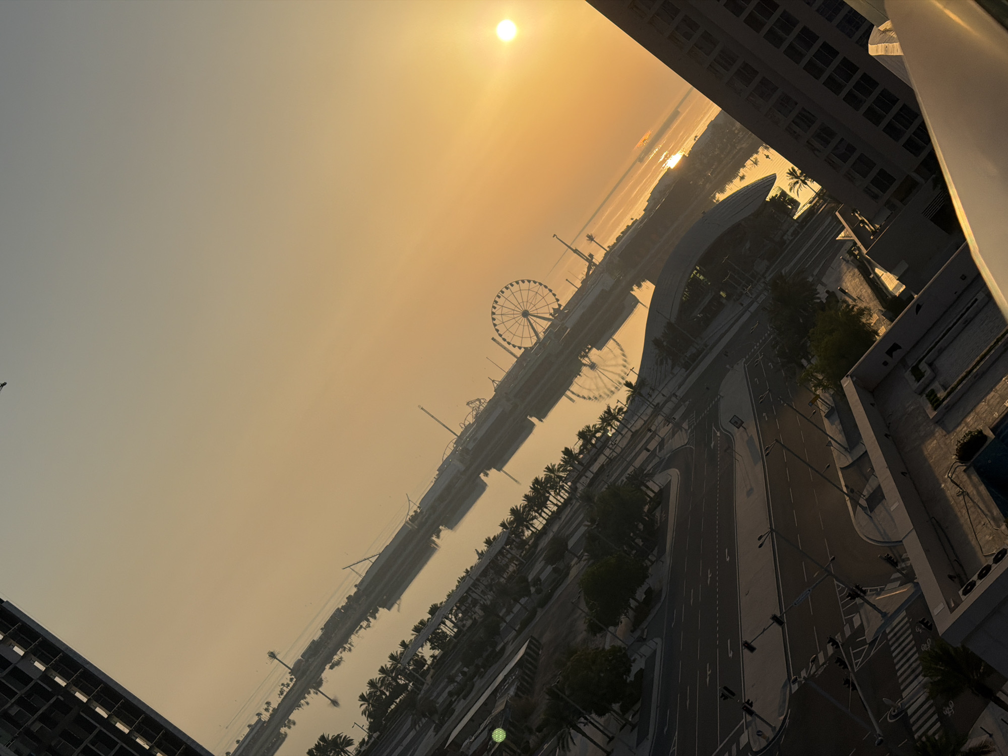 The image depicts an aerial view of a cityscape during a sunrise or sunset, with the sun visible as a bright orb reflecting on the water. The scene is dominated by a body of water, likely a harbor or bay, lined with palm trees and various structures. In the distance, a Ferris wheel stands prominently, suggesting an entertainment or amusement area. The foreground features urban infrastructure, including roads with traffic lights, pedestrian crossings, and buildings, some of which appear to be under construction or renovation. The architecture suggests a modern city with high-rise buildings. The overall atmosphere is serene, with the warm, golden light of the sun casting long shadows and creating a tranquil ambiance. The image is captured from a high vantage point, possibly a balcony or a window of a tall building, giving a sweeping perspective of the urban landscape and its reflection on the water.