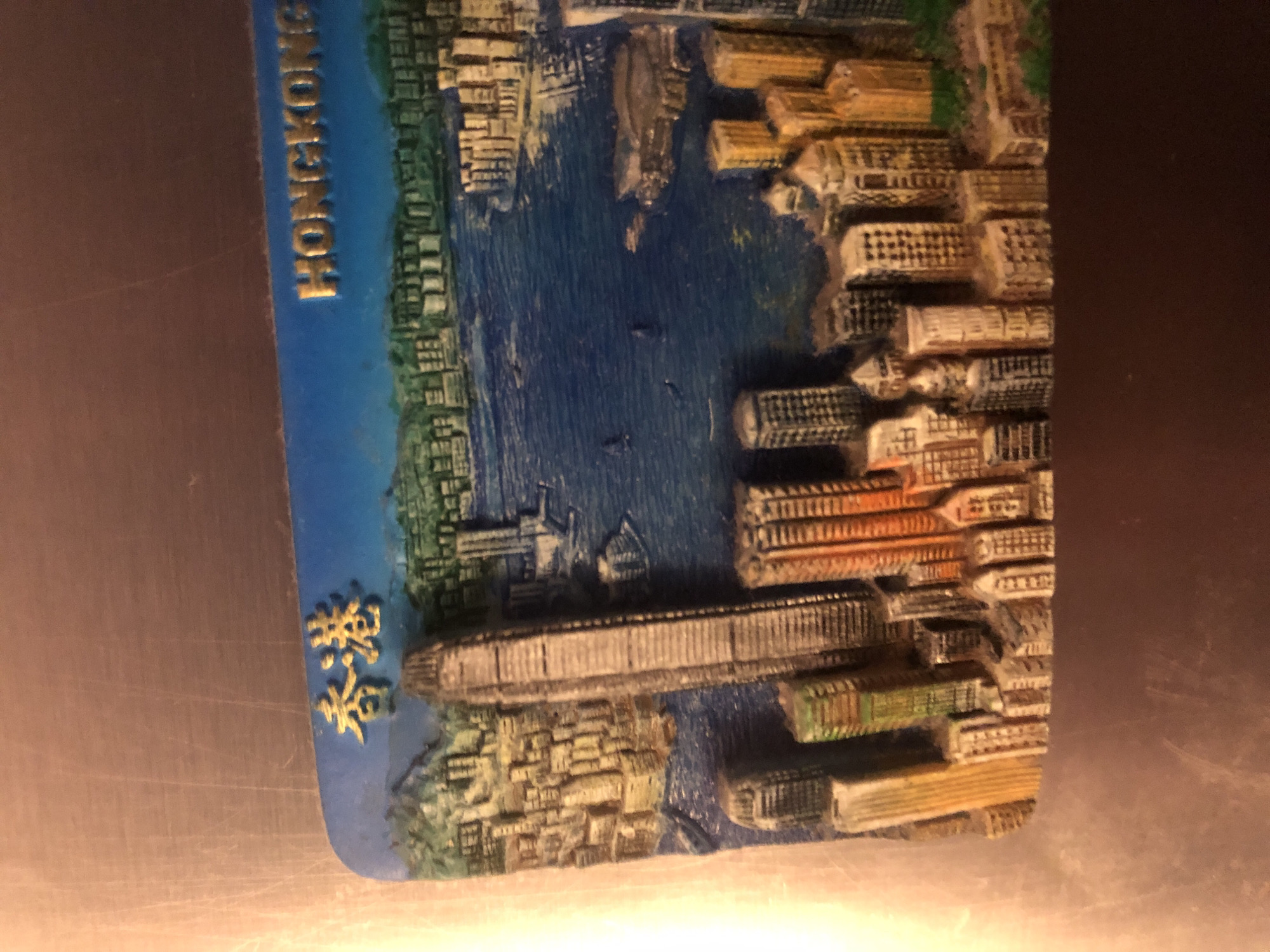 The image shows a souvenir magnet depicting the skyline of Hong Kong. The magnet features a blue backdrop representing the water and sky, with miniature buildings clustered along the coastline. Prominently displayed are several skyscrapers of varying heights and designs, including a tall, slender building that resembles the International Commerce Centre. The buildings are painted in different colors and textures, giving a sense of depth and detail. In the upper left corner of the magnet, golden Chinese characters are visible, reading "香港," which translates to Hong Kong. In the upper right corner, golden English letters spell out "HONGKONG." The magnet is placed on a metallic surface, likely a refrigerator, which has visible scratches and a brushed texture. There is no discernible connection to Arfa, Russia, as the magnet clearly represents Hong Kong.