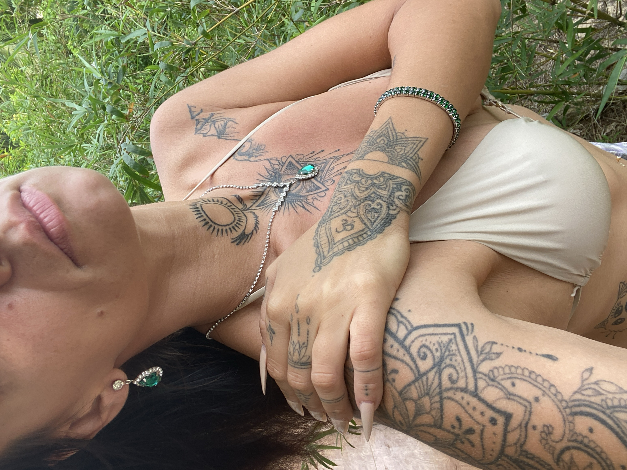 An individual, partially visible from the chest up, stands outdoors against a backdrop of dense green foliage, likely bamboo, suggesting a natural environment. The person is wearing a light-colored bikini top and accessorized with a silver necklace featuring a prominent green pendant, a matching pair of emerald drop earrings, and a tennis bracelet adorned with green stones. Their skin is extensively tattooed with intricate, dark designs, including mandalas on the arms and chest. The person's right hand is placed across their chest, revealing long, light-colored acrylic nails. Their lips are slightly parted, and their gaze is directed downwards, out of frame. The lighting suggests it is daytime, possibly late afternoon, given the soft illumination. The setting appears to be a garden or natural reserve, with the dense green growth indicative of a humid climate such as that found in Mũi Né, Vietnam, known for its tropical vegetation and coastal landscapes.