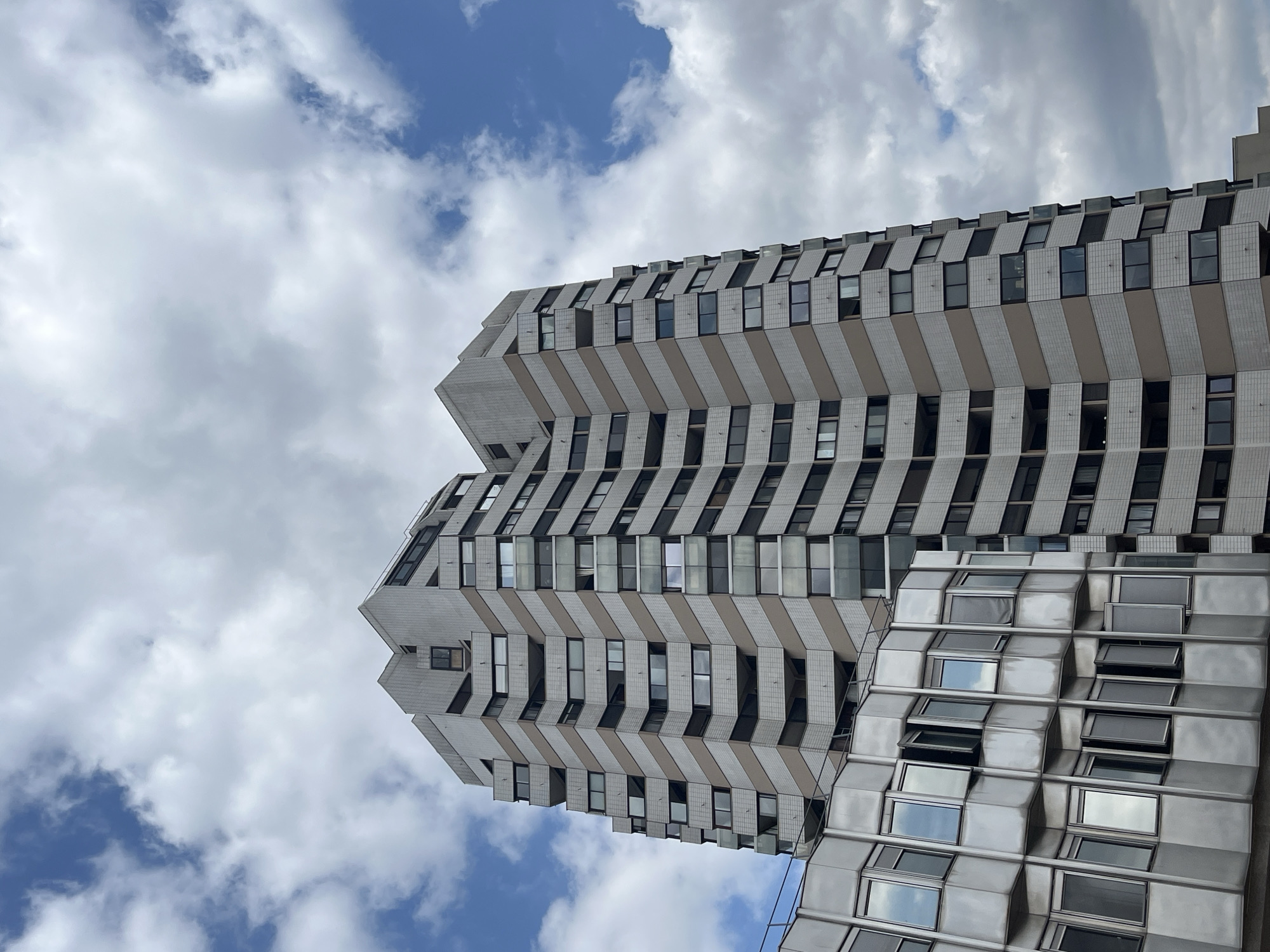 The image shows a view of the residential skyscraper known as Tour du Lac in Créteil, France, seen from a lower angle against a cloudy sky. The tower is characterized by its staggered, zig-zagging facade, which features a repeating pattern of light-colored horizontal bands alternating with darker sections containing windows and balconies. To the left of the main tower, a lower building with a more metallic, angular facade and numerous reflective windows is partially visible. The sky is a mix of blue patches and large, dramatic clouds. No people are visible in the frame. The scene depicts modern architecture in an urban environment.