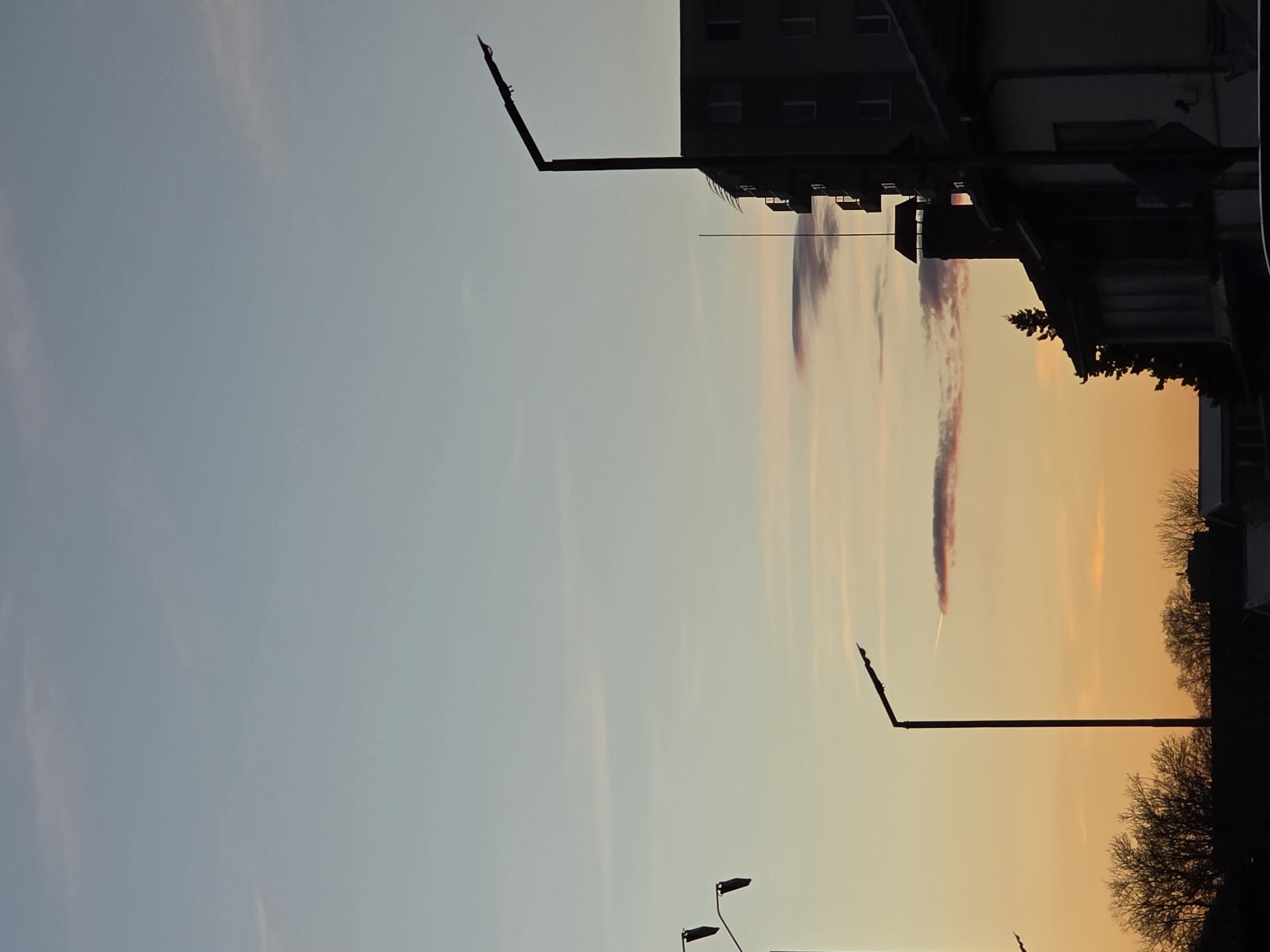 The image depicts a view in the city of Plovdiv, Bulgaria, at dusk. The sky transitions from pale blue in the upper left to orange on the right. A contrail is visible in the orange sky. Silhouetted buildings and trees are visible on the right. There are also two lamp posts on the bottom and an angled pole on top.