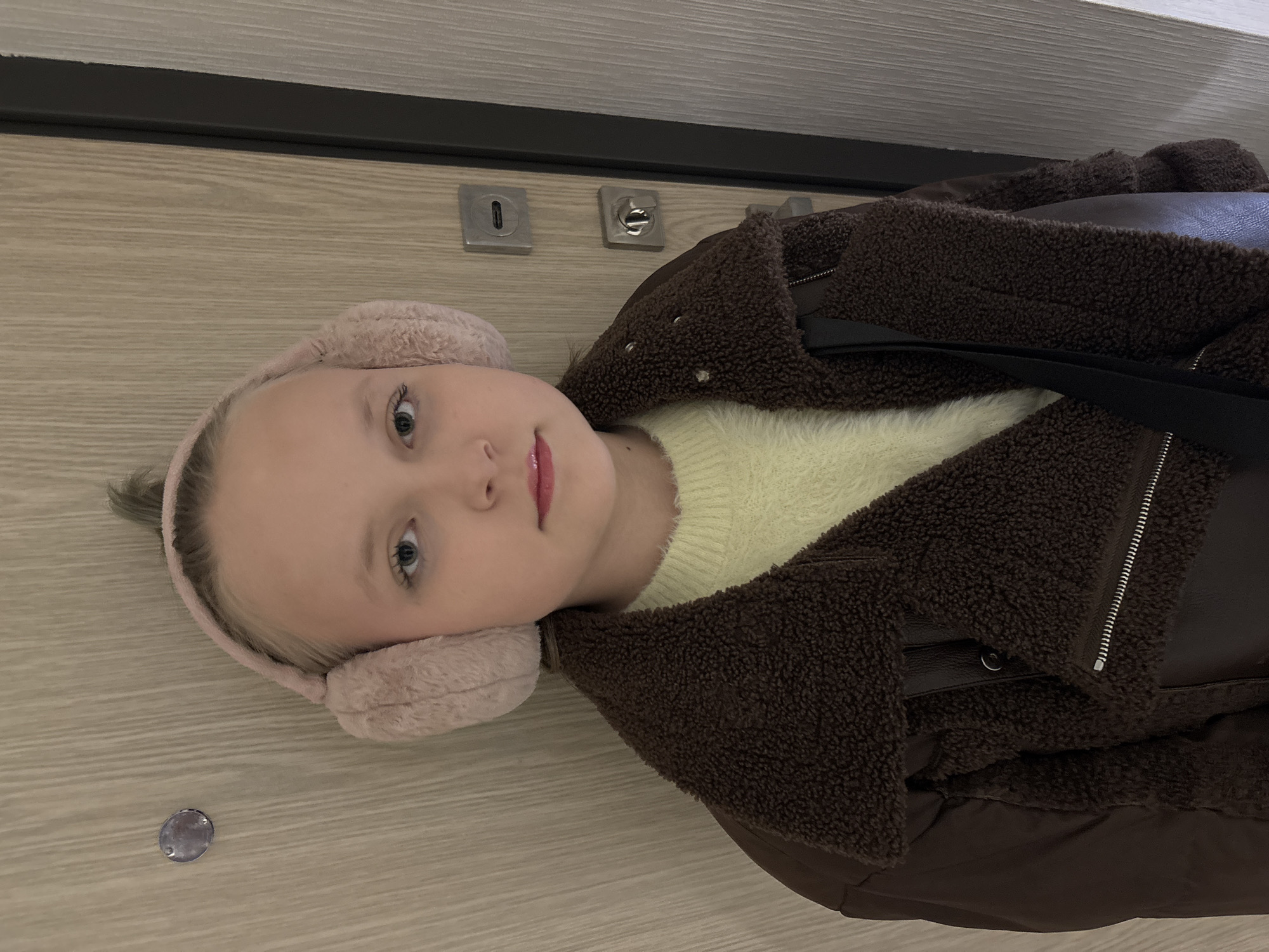 A young female with blonde hair, wearing pink fluffy earmuffs, a light yellow long-sleeved top, and a brown fleece-lined aviator jacket, is centered in the frame. A black strap from a bag is visible over her right shoulder. She looks directly forward with a neutral expression, her lips slightly parted. The background features a light brown, vertically-grained wooden door, consistent with an interior residential setting. On the right side of the door are two square, silver-toned lock mechanisms, positioned vertically. In the upper left portion of the door, a circular, translucent object, consistent with a peephole lens, is visible. The scene depicts an indoor environment. No distinct landmarks or architectural features are present to identify a precise location within Izhevsk, Russia.