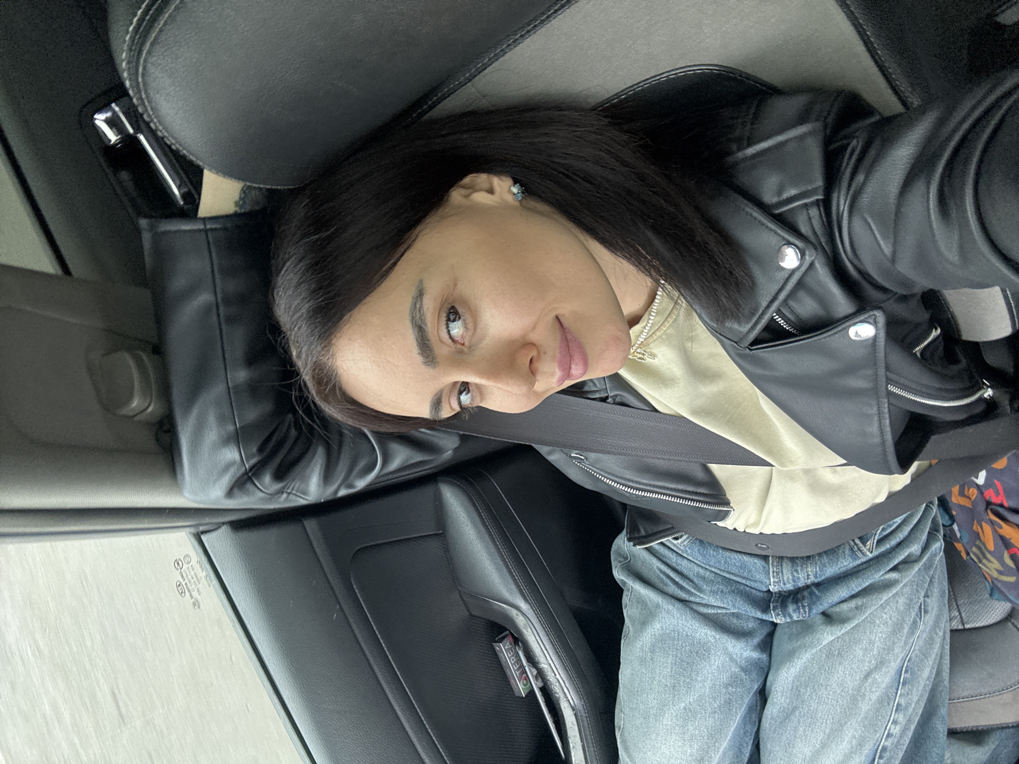 The scene depicts an adult female inside a KIA brand vehicle, likely located in Tbilisi, Georgia. She is positioned in what appears to be the front passenger seat, secured by a black seatbelt across her chest and lap. The female has dark hair, light brown eyes, and is wearing a black faux leather jacket with silver hardware over a light beige top and light blue denim jeans. A small, multi-stone stud earring adorns her right ear, and a light-colored necklace with small charms is visible. She looks directly towards the camera with a slight smile. The vehicle's interior features black and grey upholstery. A black door panel, with a textured surface and padded armrest, is visible to her left. On this armrest, a green and black package of TEREA branded tobacco sticks and a device, consistent with a heated tobacco product system, are placed. The window to her left shows a blurred, light grey background, indicative of an outdoor environment, possibly in motion or under overcast conditions.