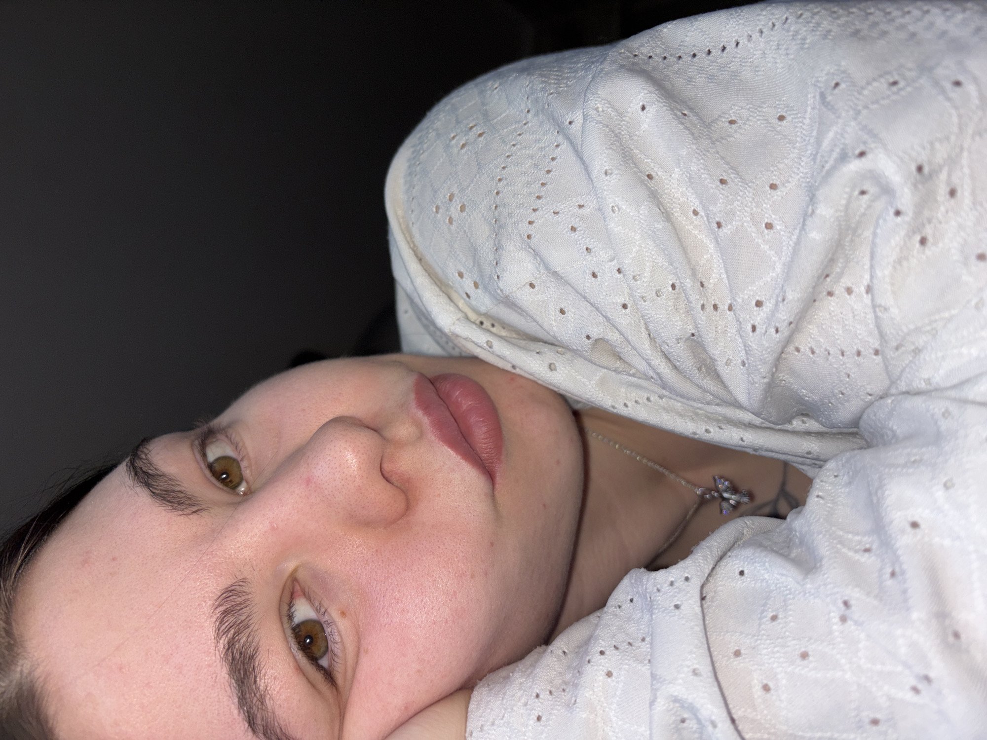 A close-up, low-angle shot depicts a person with fair skin, light brown eyes, and full lips. They are partially covered by a white, patterned garment with small perforations. A silver necklace with a butterfly pendant is visible on their neck. The background is dark and indistinct. The location in the city of Perm, Russia is not discernible from the visual information.