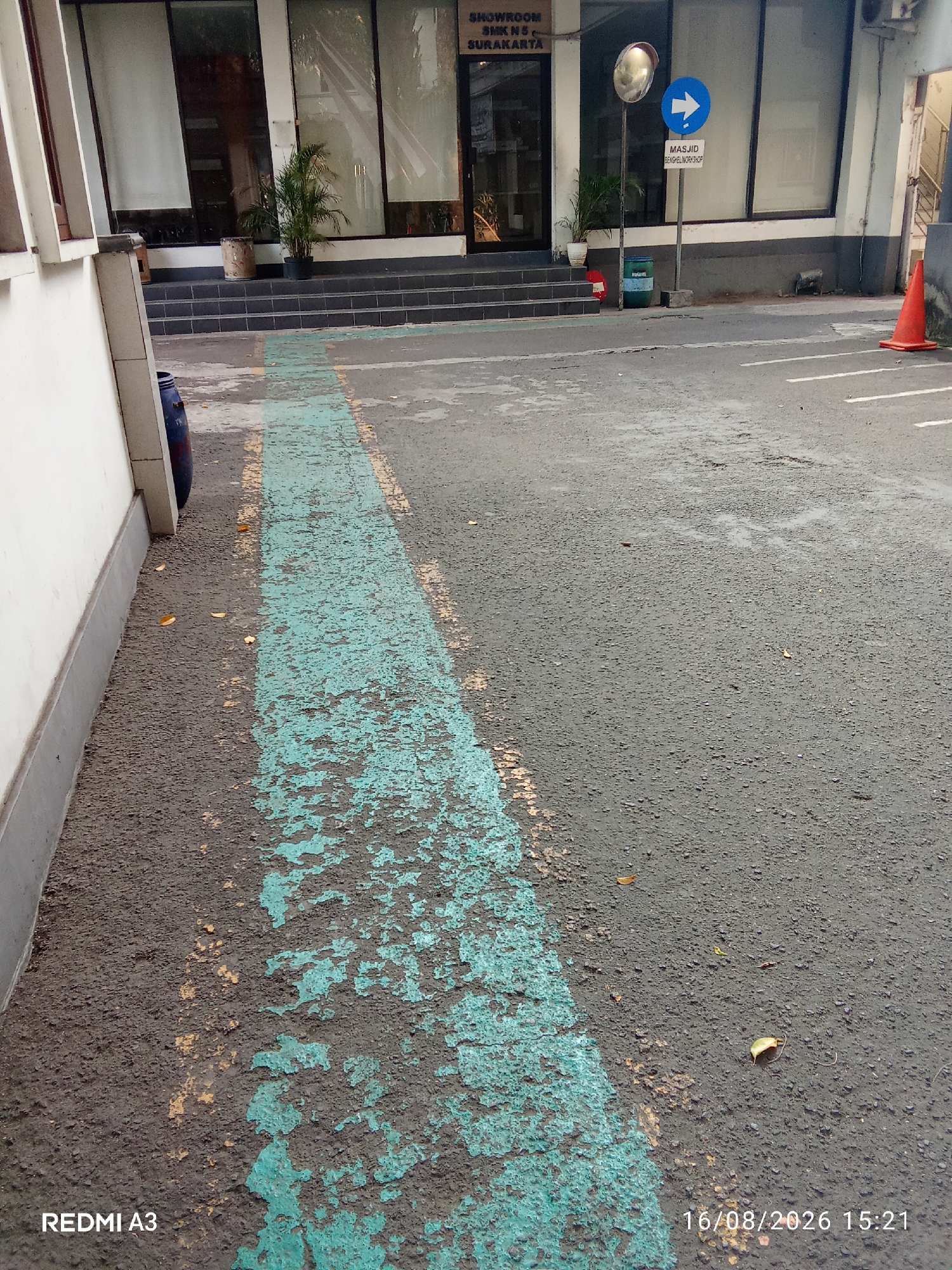 The image shows an outdoor scene on a paved surface with a faded turquoise lane marker. A white building with large glass windows and a step entrance is visible in the background. A blue barrel is positioned to the left of the building, near the white wall. A street sign with a white arrow pointing right and text indicating "MASJID BENGKEL WORKSHOP" is mounted on a pole. Further to the right, an orange traffic cone is placed on the pavement. The ground appears slightly wet in places. The timestamp at the bottom of the image reads "16/08/2026 15:21" and the text "REDMI A3" is also visible. The presence of "SURAKARTA" on the building indicates the location is in Surakarta, Indonesia.