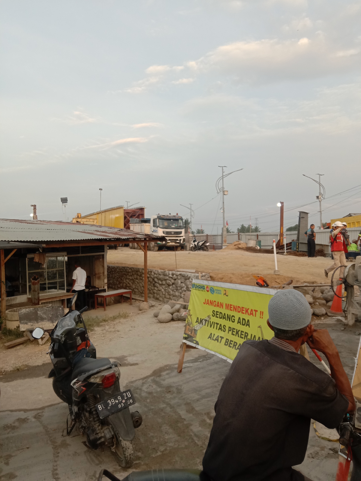 The image depicts an outdoor construction site in Lhoknga, Indonesia, at dusk. In the foreground, a person wearing a gray knit cap and dark clothing is seated on a motorcycle, facing away from the camera and resting their hand on their chin. To the left, a parked motorcycle with license plate BL 3949 ZU is visible. A small, open-sided structure with a corrugated metal roof stands to the left, with a person in a white shirt standing inside. Beyond this structure, a yellow construction beam is being transported on a truck, and a large white truck with "BSK" written on its side is parked. Further back, the site features piles of sand, construction materials, and heavy machinery, including a crane. A yellow sign with red lettering in Indonesian reads "JANGAN MENDEKAT !! SEDANG ADA AKTIVITAS PEKERJAAN ALAT BERAT" (DO NOT APPROACH!! HEAVY EQUIPMENT WORKING ACTIVITIES ARE IN PROGRESS). Several construction workers in high-visibility vests and hard hats are present on the site, with some appearing to be observing the area. The sky is overcast with faint clouds.