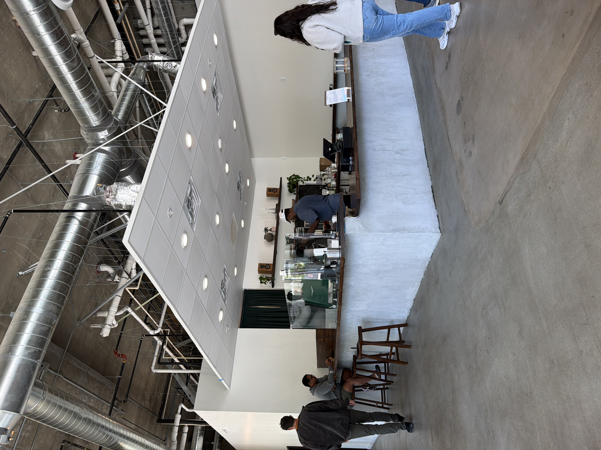 The image shows an interior shot of a modern coffee shop, possibly located in San Diego, United States of America. A woman in a light-colored sweater and blue jeans stands at the counter. Behind the counter, a barista is working, wearing a grey shirt. Two men are seated at a table nearby, one appearing to be looking at his phone. The coffee shop's interior features exposed industrial ductwork on the ceiling contrasted with a grid of recessed lights. The floor appears to be polished concrete. There are dark green curtains, shelves displaying various items, and wooden stools positioned in the seating area. The overall atmosphere is bright, minimalist, and industrial-chic.