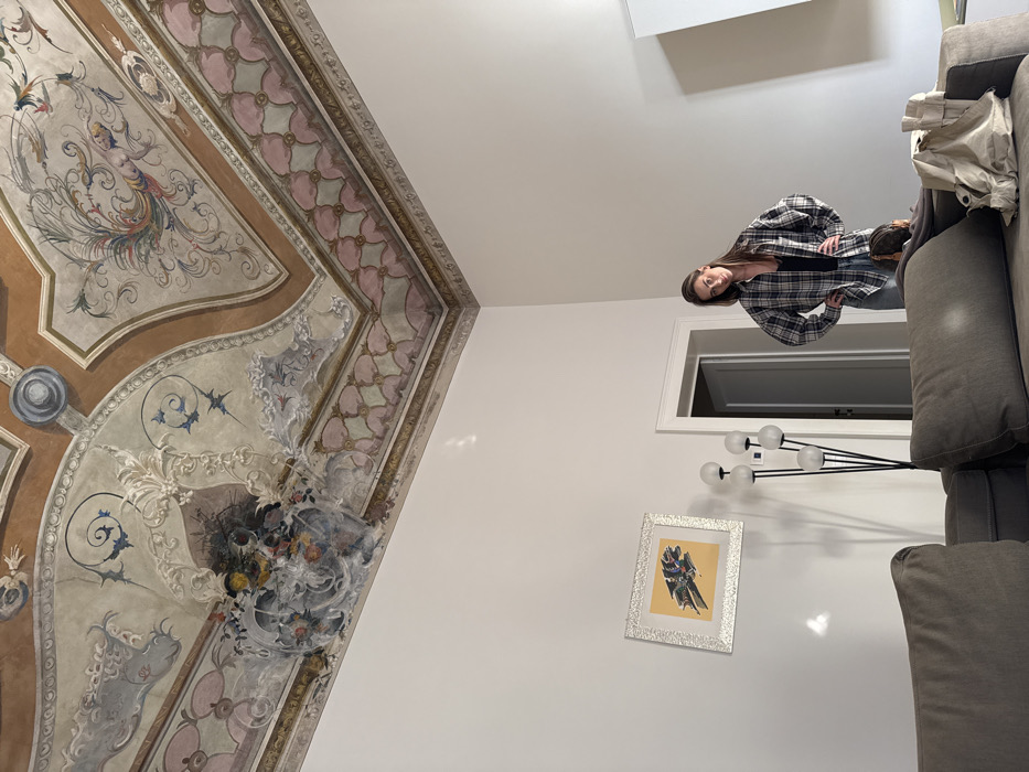 The scene depicts an interior room, likely within a historic palazzo in Venice, Italy, characterized by a high ceiling featuring elaborate frescoes. The ceiling is adorned with classical Baroque or Rococo style paintings, showcasing a central floral arrangement in a cartouche, surrounded by scrolling acanthus leaves, stylized birds, and delicate tendrils in a palette of muted pink, sage green, light blue, ochre, brown, and grey. A patterned frieze in pink and green geometric motifs borders the frescoed area.
The walls are predominantly plain white, contrasting with the ornate ceiling. On the left wall, a framed artwork with a yellow background and an abstract, multi-colored geometric design is mounted. Below it, a modern floor lamp with four white spherical lights on black stems is positioned.
In the mid-ground, a young adult female stands upright, facing the camera with her hands on her hips. She wears glasses, a loose-fitting black and white plaid button-up shirt over a dark tank top, and light-colored jeans. Her hair is long and dark.
In the foreground, a large, grey sectional sofa occupies the lower portion of the image. Behind the sofa, to the right of the standing female, the top of a head with brown hair belonging to a second individual, possibly a child, is partially visible. A light-colored canvas bag or backpack rests on the sofa to the right of the partially visible person. A white doorway is visible in the background between the floor lamp and the standing female.