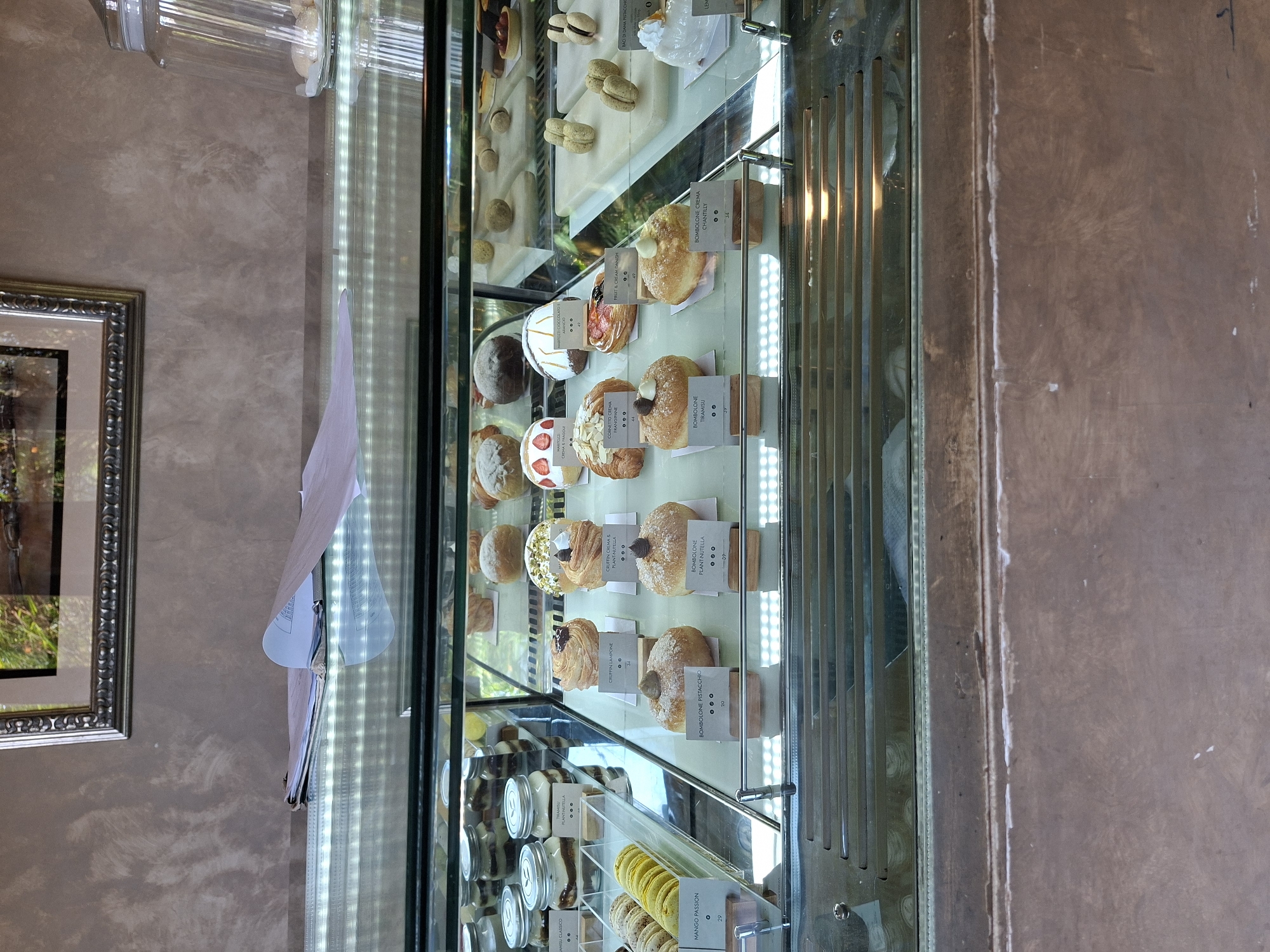 An indoor scene in what appears to be a bakery or cafe in the Ubud District, Indonesia, displays a brightly lit glass pastry case filled with a variety of desserts. The image is oriented vertically but depicts the display case as if viewed front-on.
The main subject is a multi-tiered glass display case, illuminated by vertical LED strips, showcasing numerous baked goods and sweet treats. On the upper shelves, various pastries are arranged, including several types of bombolone donuts, identifiable by their round, filled appearance and labels such as "BOMBOLONE PISTACCHIO," "BOMBOLONE PLANT-NUTELLA," "BOMBOLONE TIRAMISU," and "BOMBOLONE CREMA CHANTILLY," with visible prices like "20" and "29." There are also croissants and danishes, including "CORNETTO CREMA FRANGIPANE," "CRUFFIN CREMA &" (likely Frangipane or Fragole), "CRUFFIN LAMPONE," and a "FRUIT & CREAM DANISH." Small white labels with black text identify each item.
The lower shelves feature jars containing layered desserts, explicitly labeled "TIRAMISU CLASSICO" and "TIRAMISU DELLA" (presumably a variation), alongside macarons in flavors like "MANGO PASSION" and "BACI DI DAMA PISTACCHIC." Some macarons appear to be light green, yellow, or off-white.
In the background, a textured, light brown wall is visible, with a framed picture depicting a landscape or nature scene on the left. A clear glass jar, possibly holding more treats or ingredients, sits atop the display case on the upper left. A piece of paper or menu is attached to the side of the display case. The overall presentation suggests a clean, inviting establishment with a focus on fresh, appealing desserts, possibly with an Italian influence given the names of many items.